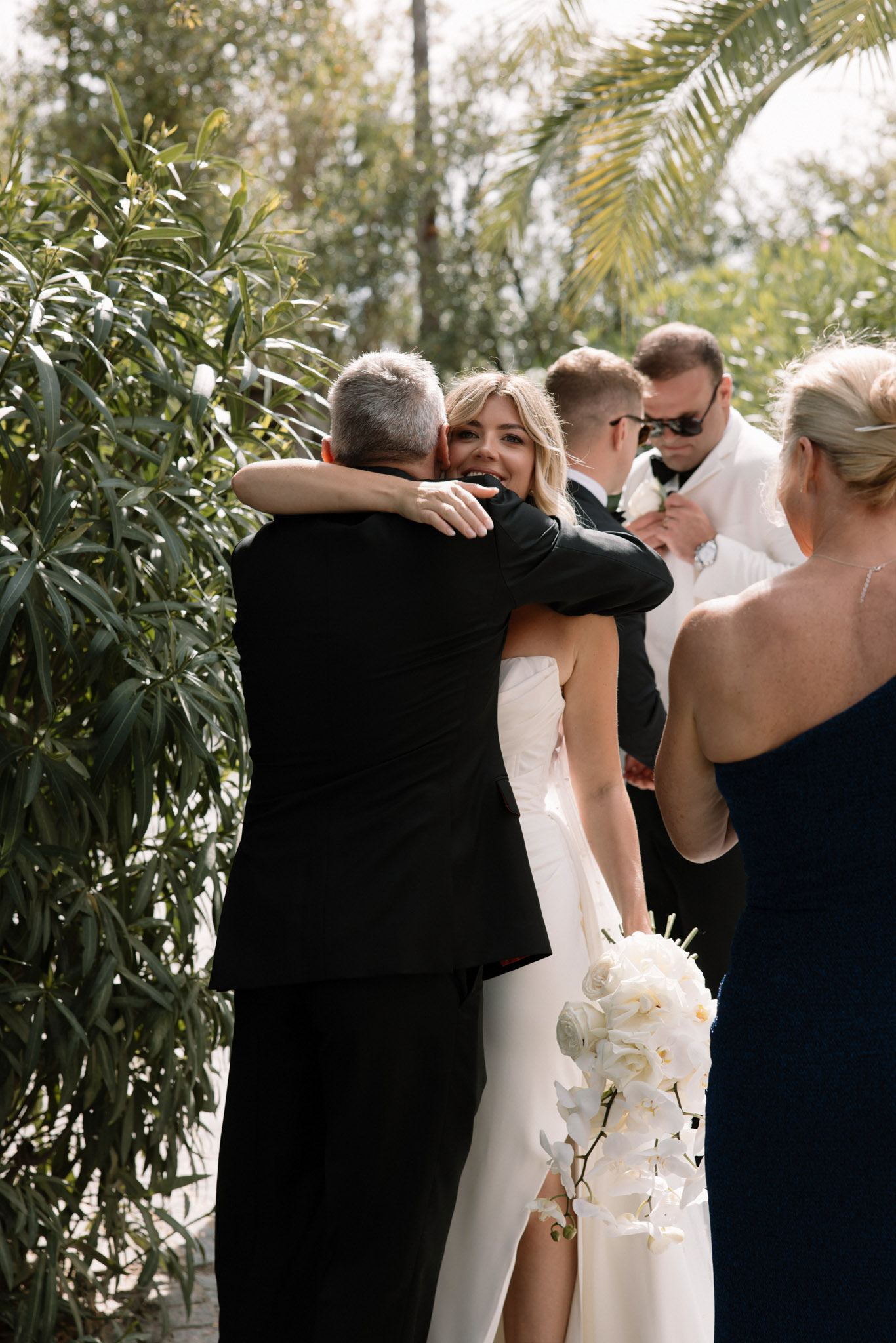 The bride embraces a male guest in a black suit during what appears to be a pre-ceremony or post-ceremony moment outdoors, smiling warmly over his shoulder toward the camera. She is wearing a strapless white fitted gown with a front slit and carries a white bouquet composed of garden roses and cascading white orchids. In the background, a man in a white jacket — likely the groom — is having a boutonniere pinned to his lapel by a woman in a navy one-shoulder dress, while another male guest in a black suit stands nearby. The setting is an outdoor garden or terrace with lush greenery and palm fronds visible. The overall styling is modern and classic, with a monochromatic white and navy palette. This is a candid mid-shot capturing a genuine moment among the wedding party.
