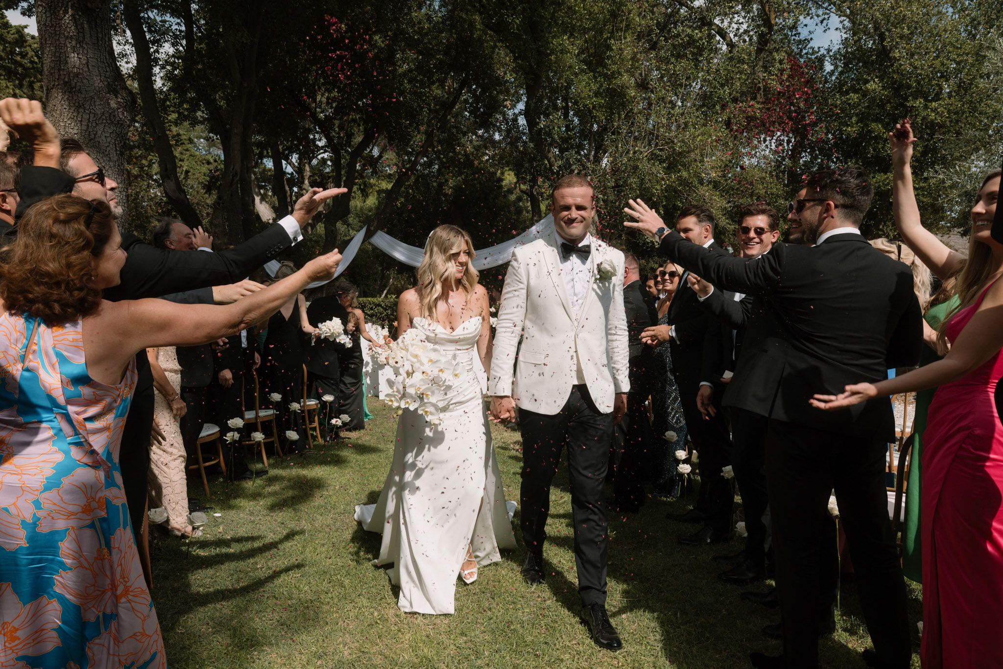 Bride and groom walk through pink petal confetti shower after outdoor garden ceremony lined by cheering guests