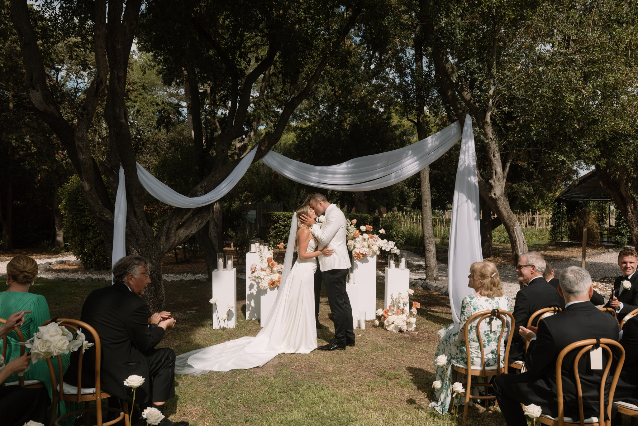 First kiss under draped white fabric with ivory and peach rose pedestal arrangements and 15 guests