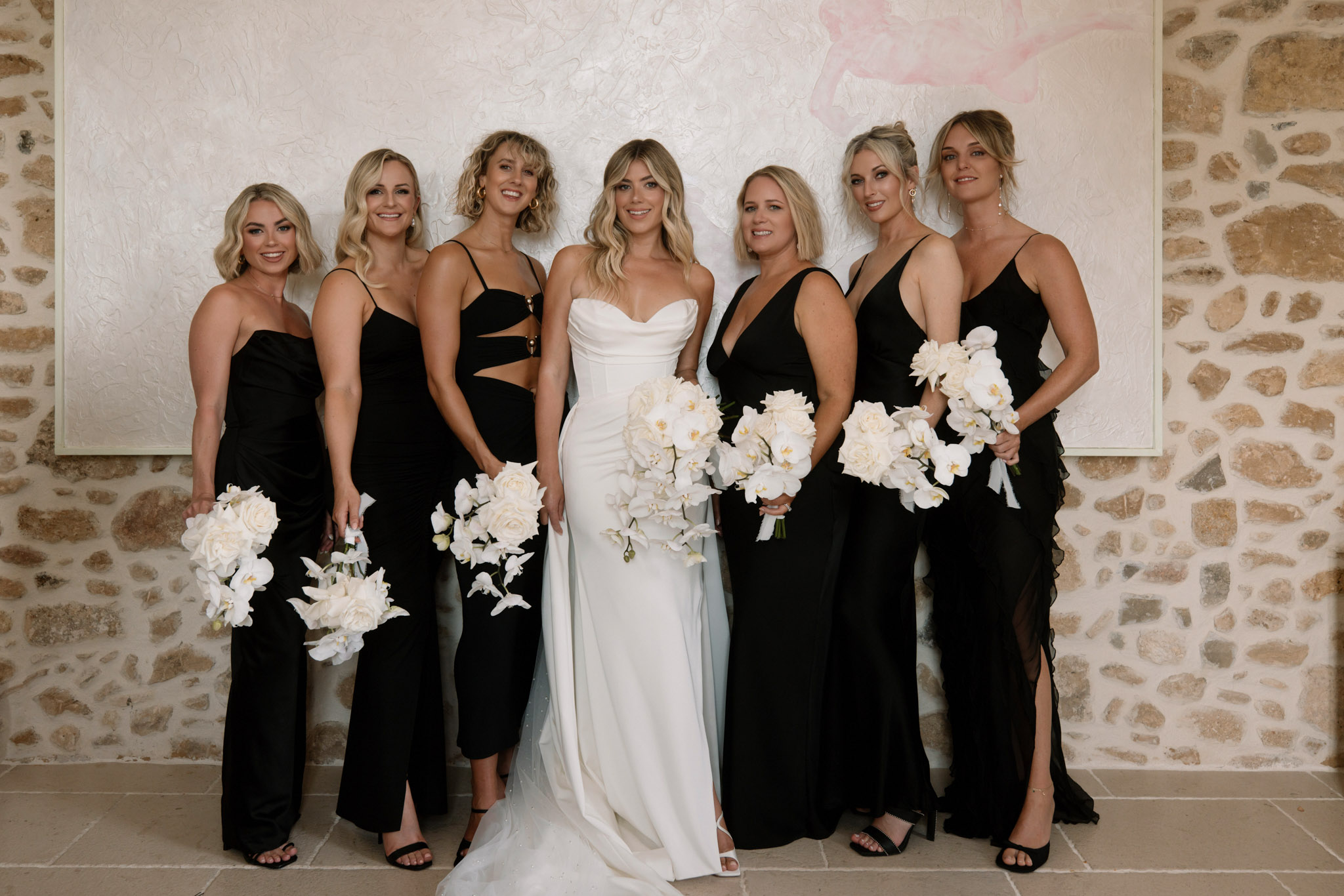 A bridal party portrait featuring the bride and six bridesmaids posed indoors against a textured plaster wall hung with a large abstract artwork in soft pink and white tones, flanked by exposed stone walls. The bride wears an ivory satin sweetheart-neckline fitted gown with a train and holds a large cascading bouquet of white phalaenopsis orchids and white roses. The six bridesmaids wear mismatched black floor-length gowns in varying styles — including spaghetti straps, cut-out details, deep V-necks, and a ruffled hem — each holding a round bouquet of white roses and white orchids. The overall styling palette is a high-contrast black and white with a modern, sleek aesthetic. This is a medium group portrait shot taken at eye level.