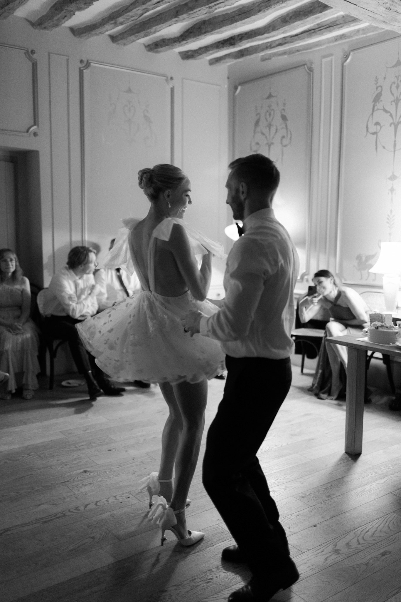 A black-and-white image of a couple sharing their first dance in an indoor reception room. The bride wears a short, voluminous mini dress with a low open back, bow shoulder detail, and embroidered or textured skirt, paired with heeled sandals with bow ankle ties and her hair in an updo; the groom is dressed in a white dress shirt and dark trousers. The room features exposed wooden ceiling beams, white panelled walls with painted decorative motifs, and wide-plank hardwood flooring, consistent with a French château or manor interior. Several guests are visible seated along the walls in the background, watching the dance, with a lamp and side table visible to the right. The image is shot from a medium-wide angle, capturing both the couple's full figures and the surrounding environment, with strong contrast between highlights and deep shadows.