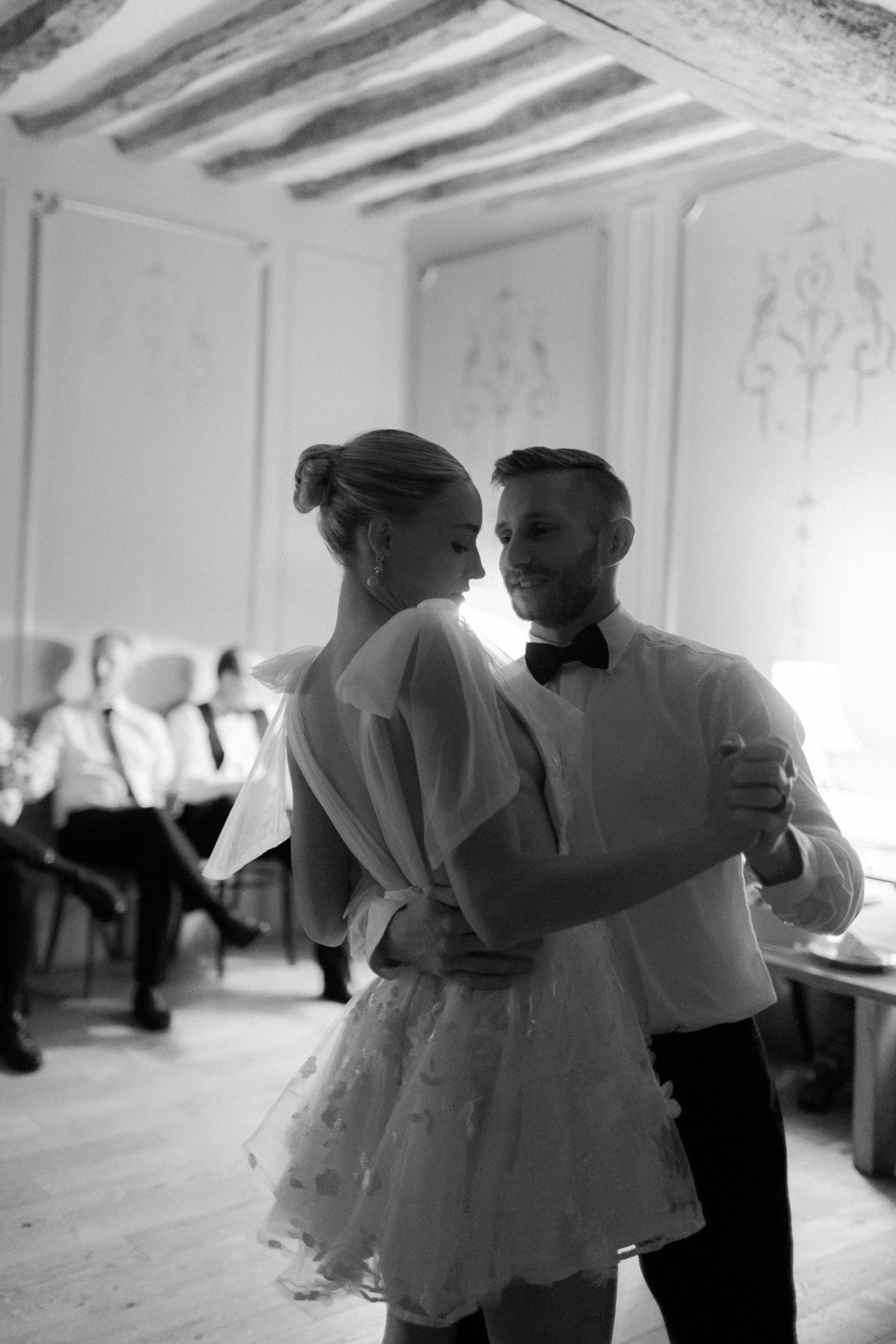 This black-and-white image captures the couple's first dance inside a classic French interior room featuring exposed ceiling beams and ornate wall paneling with decorative moldings. The bride wears a short, voluminous dress with a deeply open back, billowing sleeves, and a textured ruffled skirt, paired with her hair in an updo and drop earrings; the groom wears a white dress shirt with a black bow tie and dark trousers. The couple faces each other closely, the groom smiling, while several seated guests in formal attire are visible in the soft background. The image is shot in a medium portrait style with high contrast and bright backlighting from a window behind the couple, creating a soft glow around them.