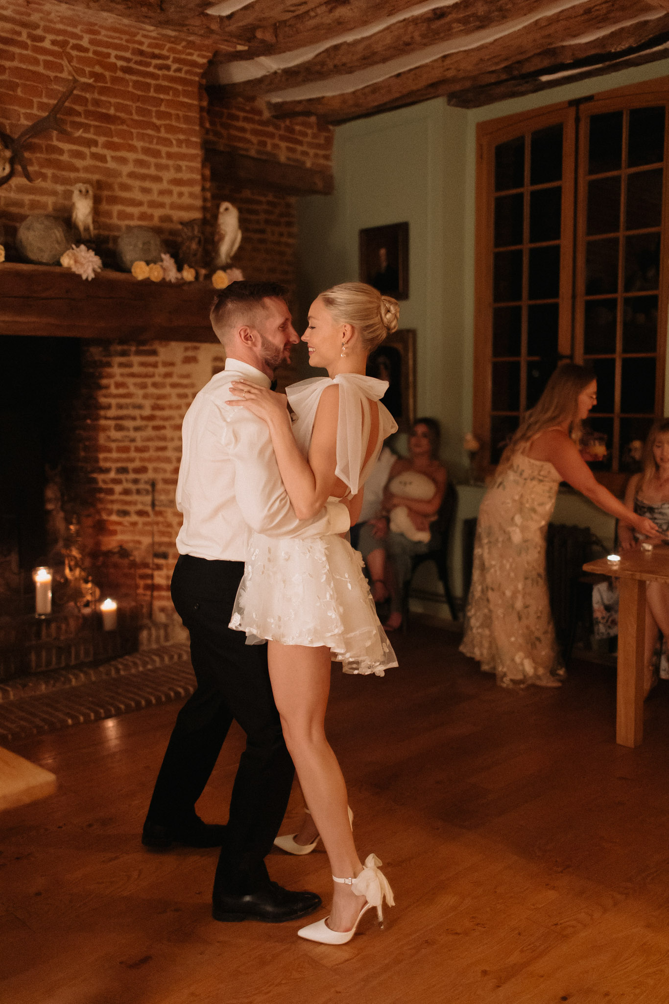 First dance in candlelit room with exposed brick and stone fireplace, bride in ivory mini dress with bows