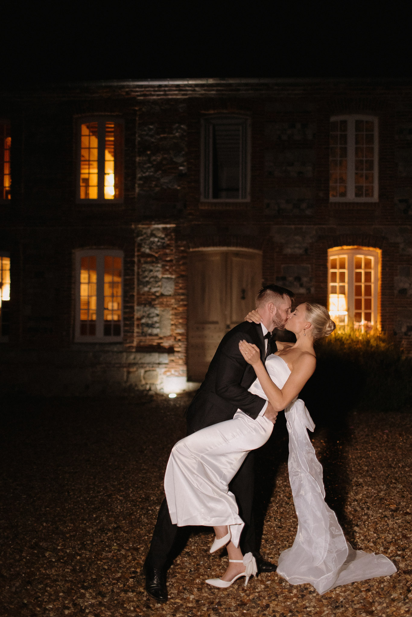 A nighttime couple portrait taken outdoors on a gravel courtyard in front of a French stone manor or château. The groom, wearing a black tuxedo with a bow tie, dips the bride as they kiss. The bride wears a strapless ivory satin column dress with a large oversized bow detail at the back and white ankle-strap heeled shoes, her blonde hair styled in an updo. The stone building behind them is warmly lit from within, with golden light glowing through multiple French windows and a rounded wooden door, and ground-level uplighting illuminating the facade. The overall styling is modern and minimal. Full-length portrait shot.
