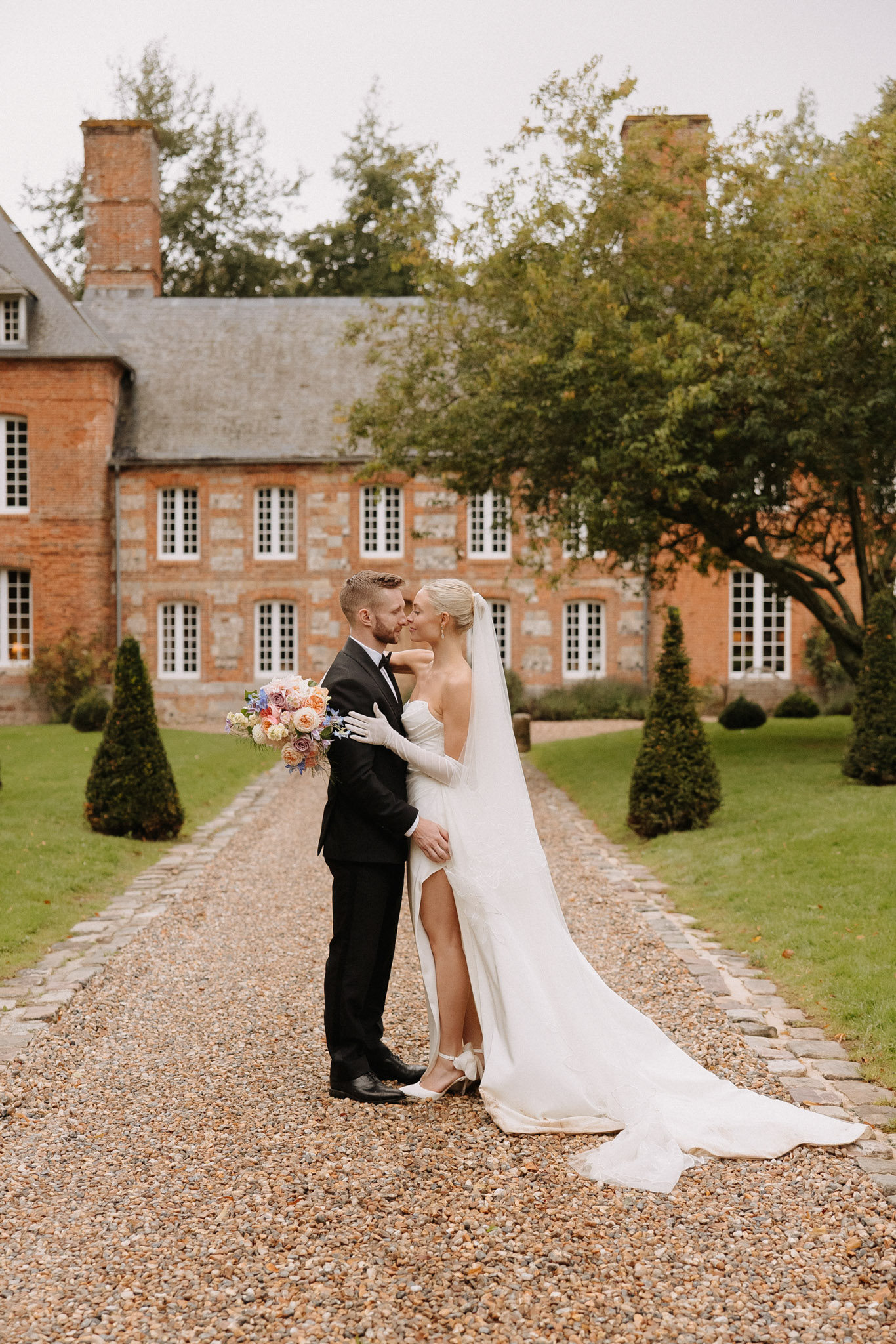 Bride in column gown with cathedral veil and white gloves kissing groom on path to red brick manor
