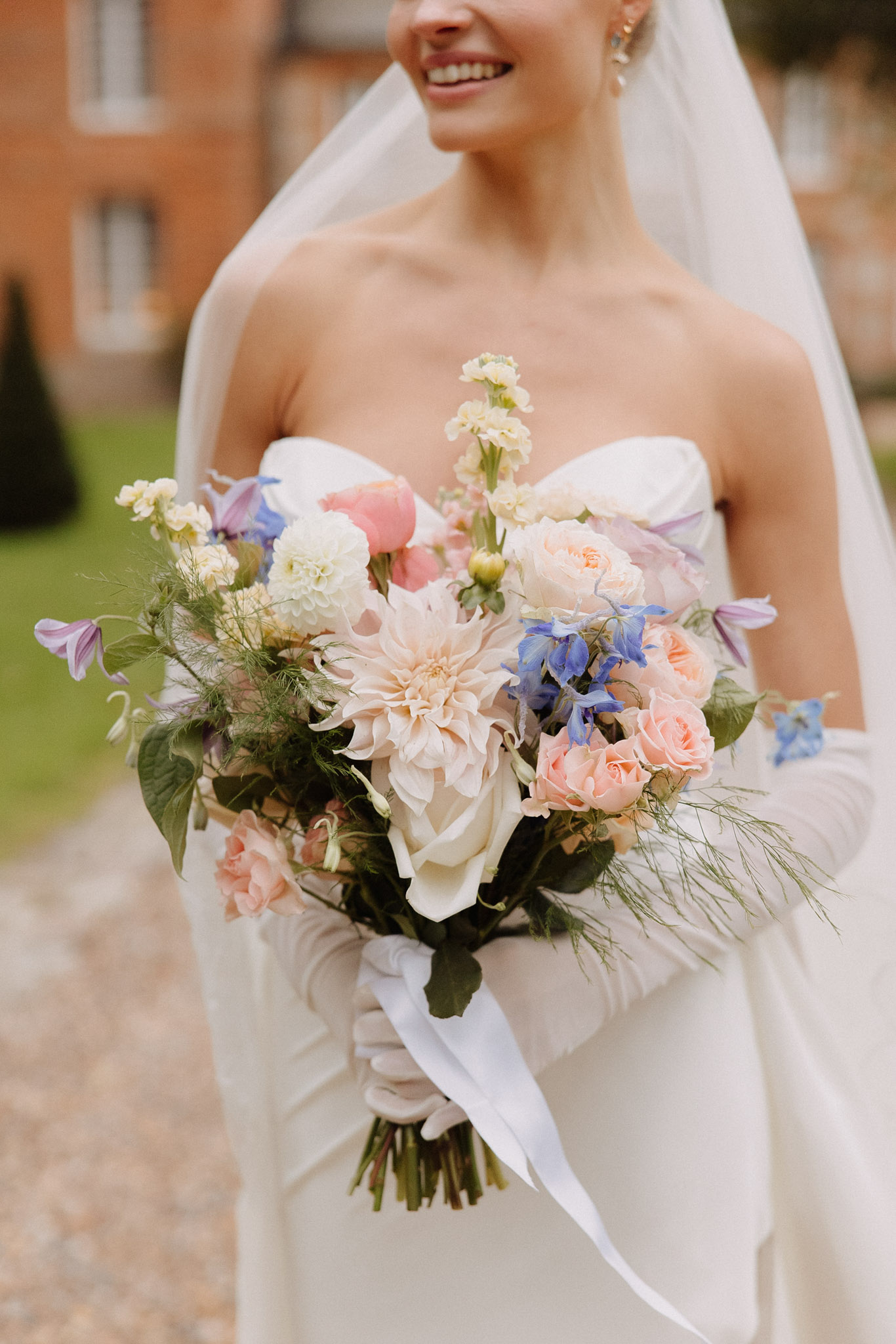 A close-up portrait of a bride outdoors on a gravel path, photographed from the chin down with a red-brick château building softly blurred in the background. She wears a strapless ivory fitted gown, long white satin gloves, and a cathedral-length veil. She holds a loose, garden-style bridal bouquet composed of blush and peach garden roses, a large café au lait dahlia, a white dahlia, coral peonies, blue delphinium, lavender clematis, cream stock flowers, white calla lilies, and trailing feathery green foliage, tied with a long trailing white silk ribbon. The bouquet palette combines soft peach, blush, ivory, cornflower blue, and lilac tones in an unstructured, botanical arrangement. The composition is a medium close-up focused on the bouquet and torso.