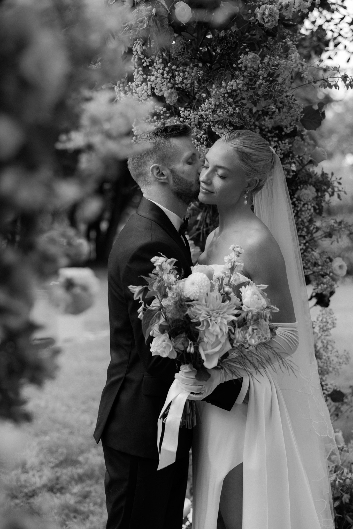 A black-and-white couples portrait taken outdoors, showing the groom kissing the bride on the temple while she smiles with eyes closed. The groom wears a dark tuxedo with a bow tie, and the bride wears a strapless or off-the-shoulder gown with long white gloves and a flowing veil. She holds a large, loosely arranged bouquet featuring dahlias, roses, and what appears to be delicate filler florals, tied with long ribbon streamers. The couple stands in front of a lush floral arch densely filled with mixed blooms and foliage. The image is shot in high contrast black and white with a shallow depth of field, rendered as a close-up portrait with soft bokeh in the foreground and background.