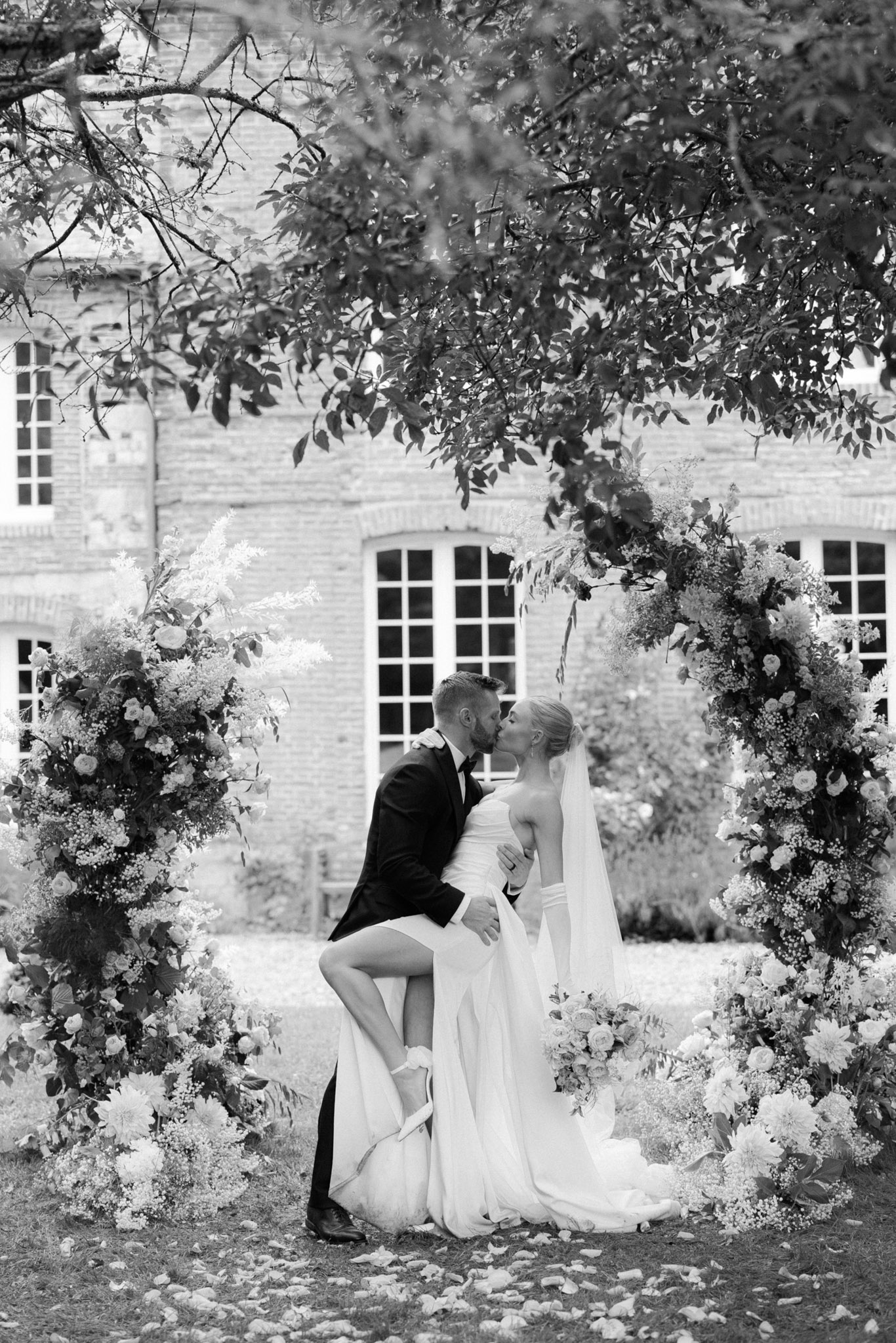 Black and white photo of bride and groom sharing dip kiss in front of circular floral arch at chateau