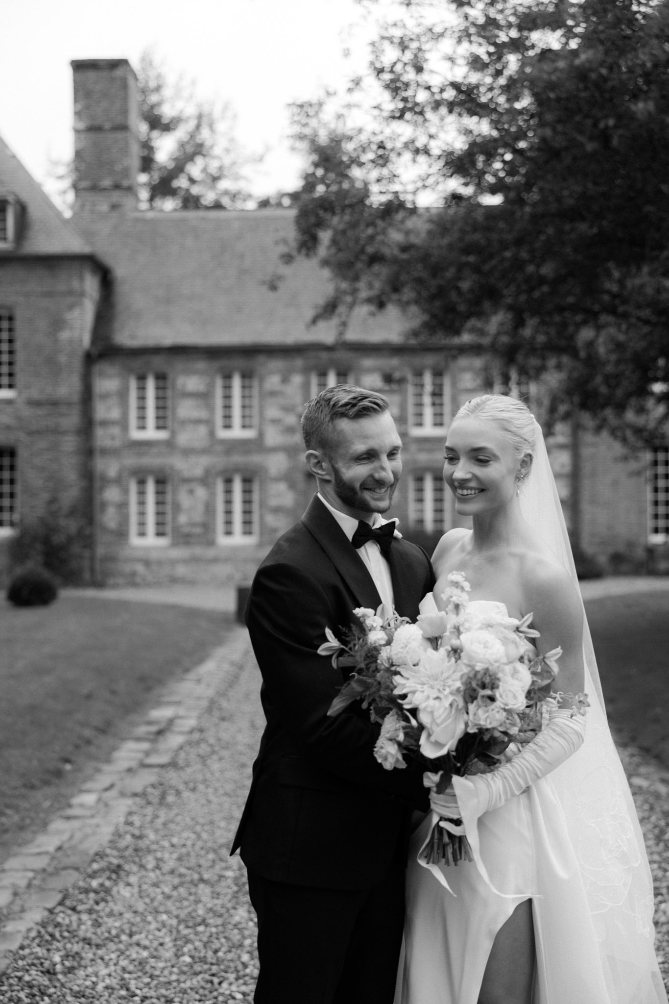 Black-and-white portrait of bride and groom laughing outside a stone manor, bride in strapless gown with long gloves and veil