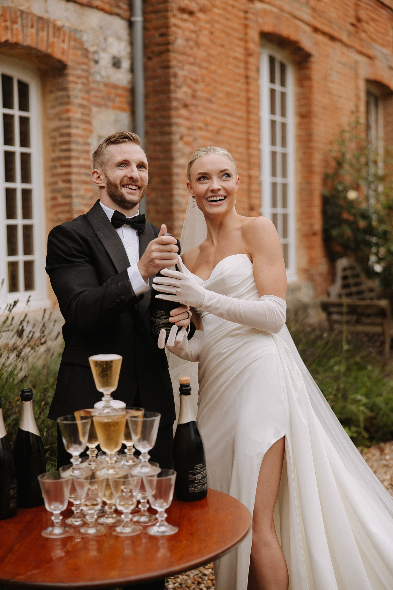 Couple popping champagne beside coupe glass tower on red brick chateau terrace