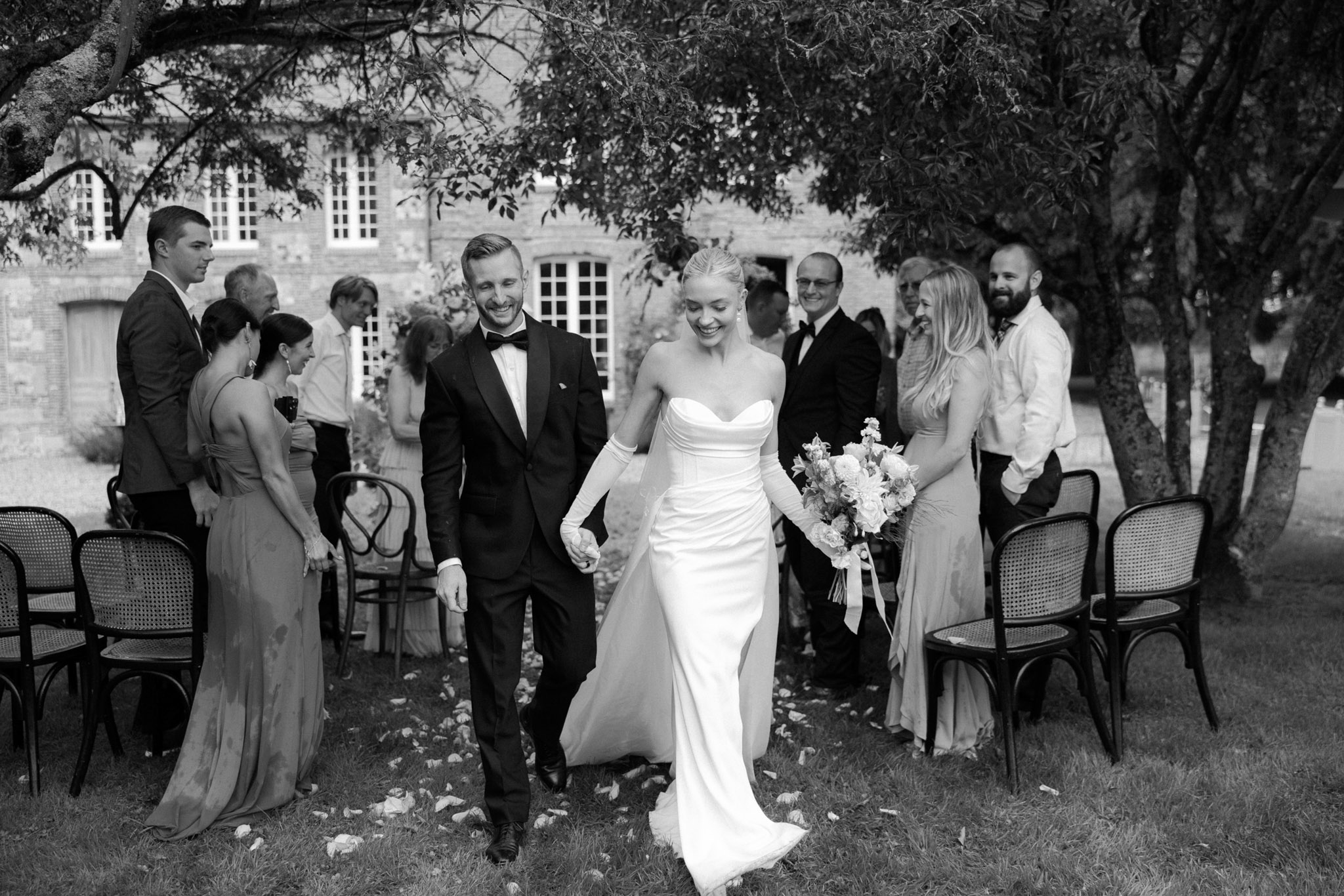 Black-and-white image of bride and groom walking back up the aisle after outdoor ceremony at a stone chateau