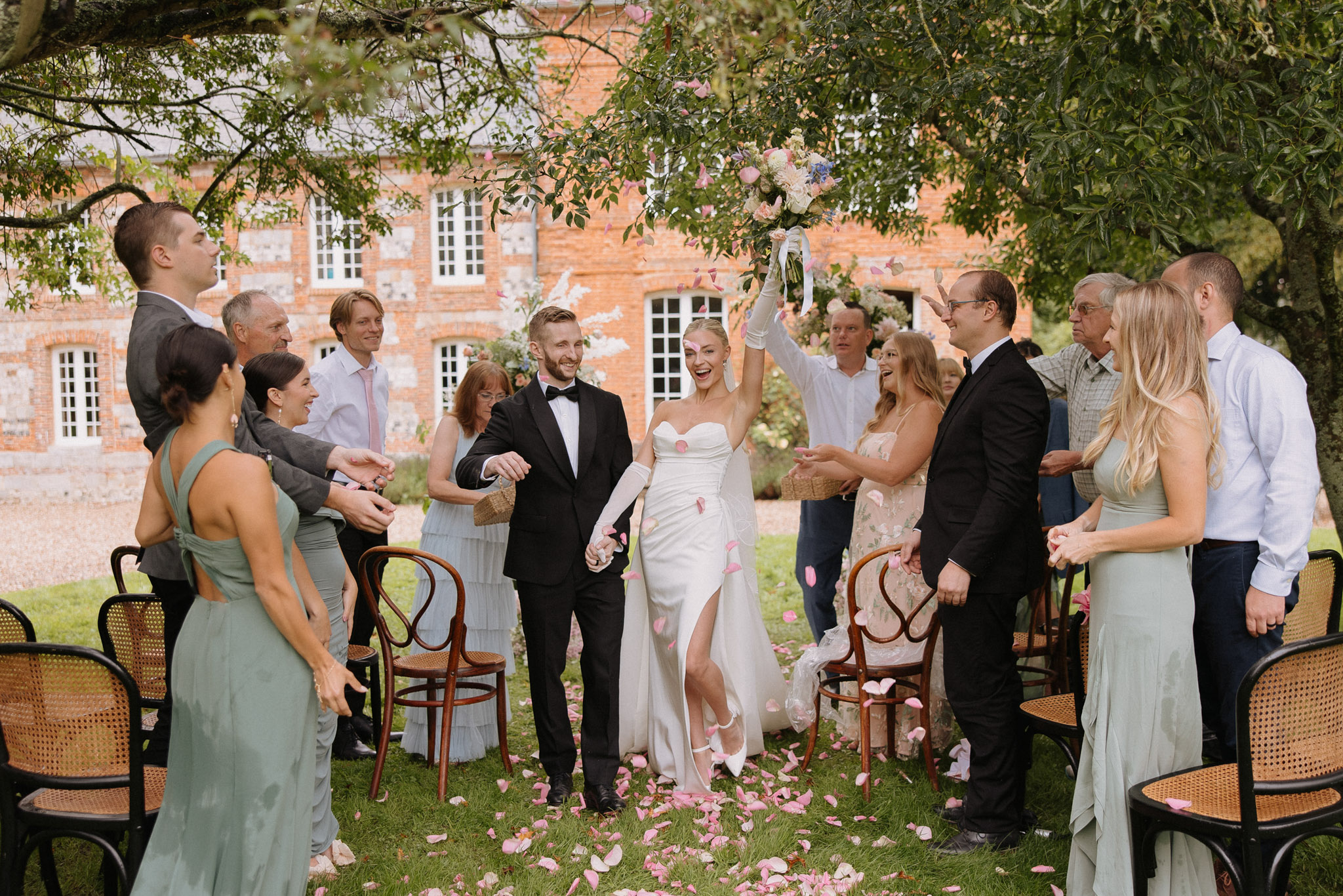 The bride and groom are walking back up the aisle as a newly married couple during an outdoor ceremony recessional at what appears to be a French brick manor house. The bride wears a white strapless fitted gown with a front slit, long white gloves, and white pointed-toe heels, and raises her mixed bouquet of cream, pink, and blue flowers above her head in celebration; the groom wears a black tuxedo with a bow tie. Guests lining the aisle — approximately 12–14 visible — are throwing pink rose petals, which are scattered across the grass aisle, and several bridesmaids wear sage green halter-neck dresses. Seating consists of classic dark bentwood café-style chairs with rattan seats, and the overall styling is modern and clean with a soft pink and sage green color palette. Wide editorial shot taken at ground level capturing the full width of the aisle and the joyful expressions of the couple and guests.