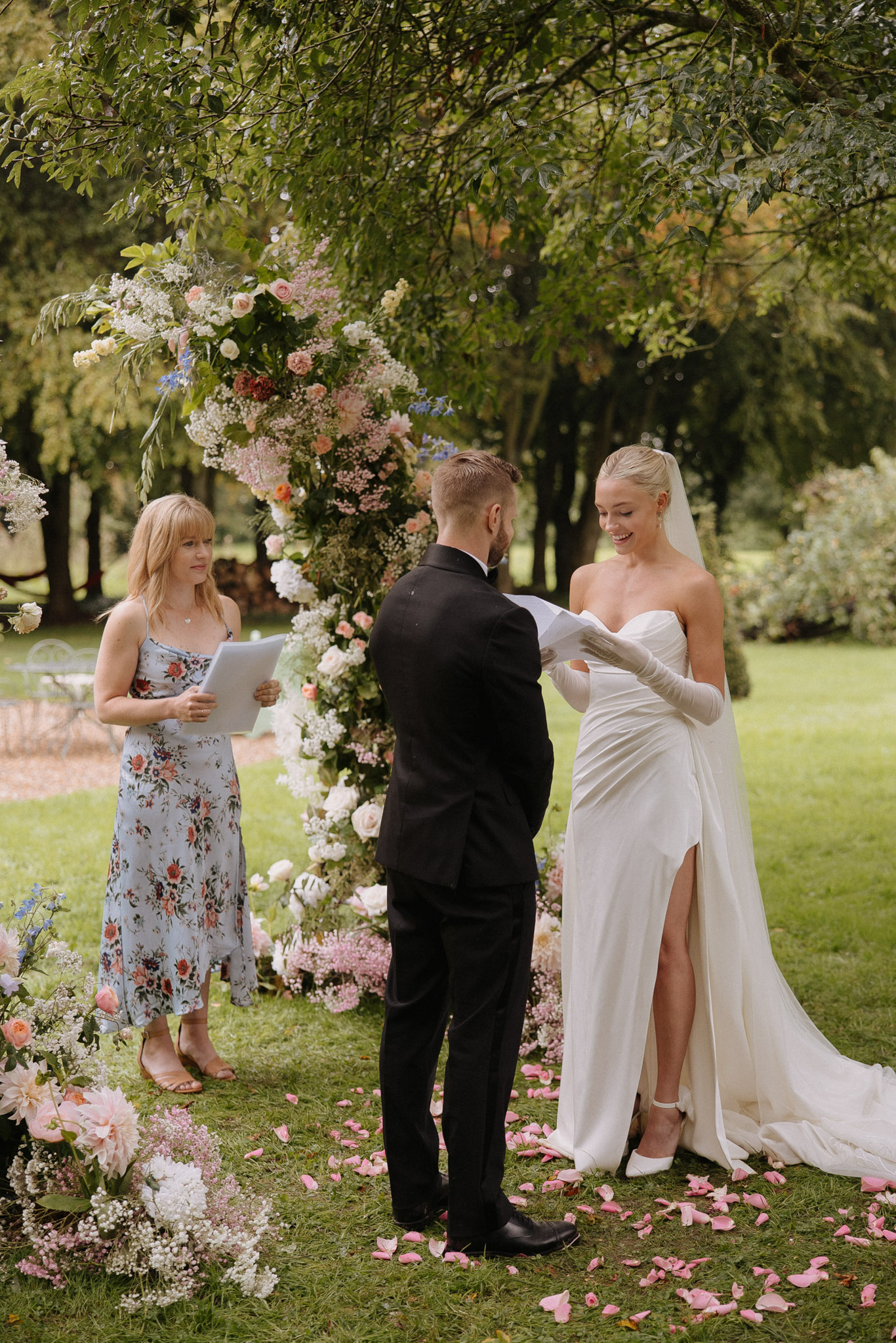 Bride reading vows to groom during outdoor garden ceremony beside a tall floral column of pink peach and blue blooms