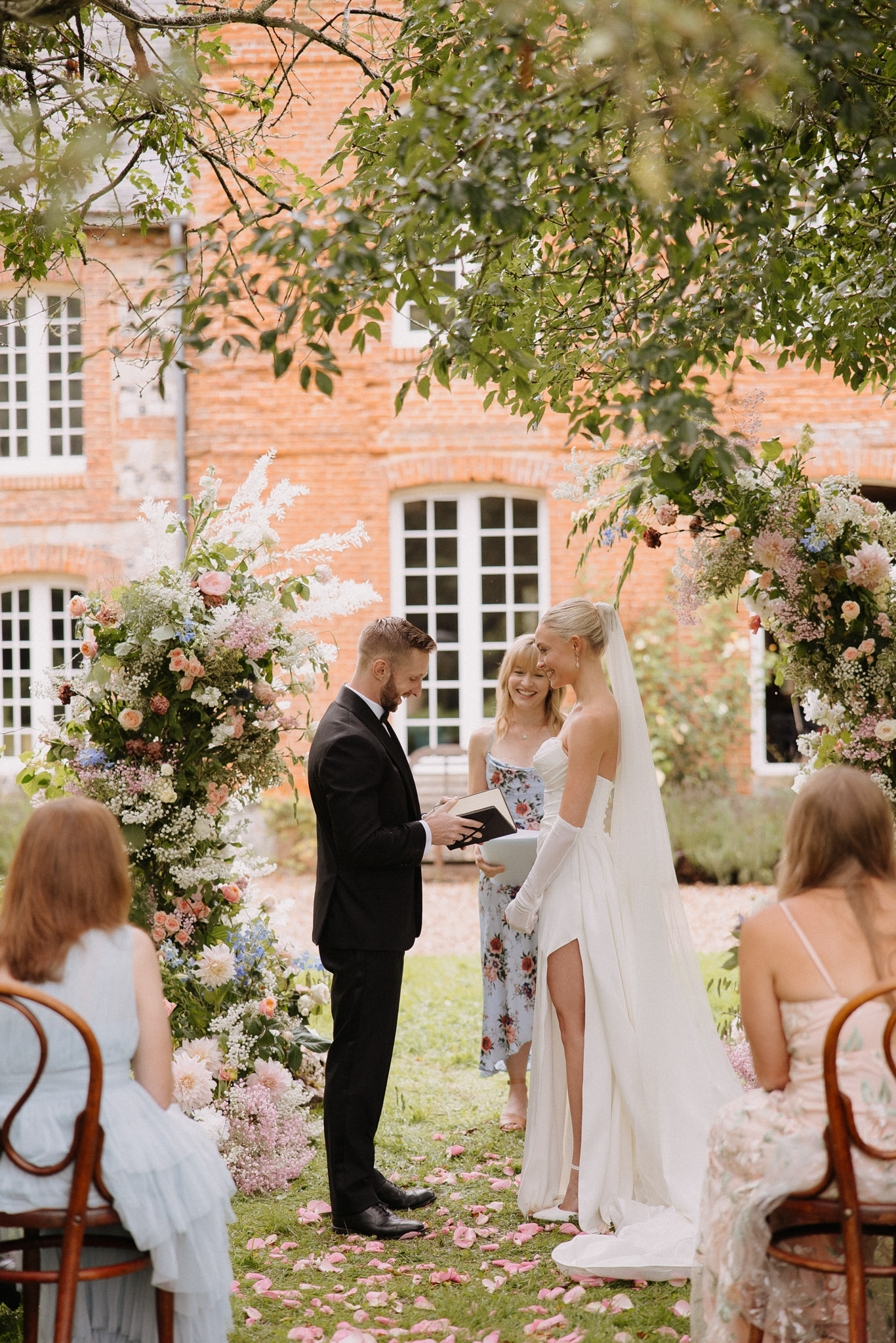 An outdoor wedding ceremony taking place in the garden of a red-brick French manor house with white-framed windows. The groom, wearing a black tuxedo, reads from a book while the bride, dressed in a fitted off-the-shoulder white gown with a long veil and long white gloves, smiles beside him; an officiant in a blue and white floral dress stands between them holding a book. Two large floral installations flank the couple, featuring blush peach garden roses, white astilbe, blue delphinium, pink dahlias, lilac, and lush greenery in a garden-style, loose arrangement. Pink rose petals are scattered along the grass aisle, and seated guests in bentwood chairs are visible in the foreground on both sides. The shot is a medium-wide portrait framed by overhanging tree branches.