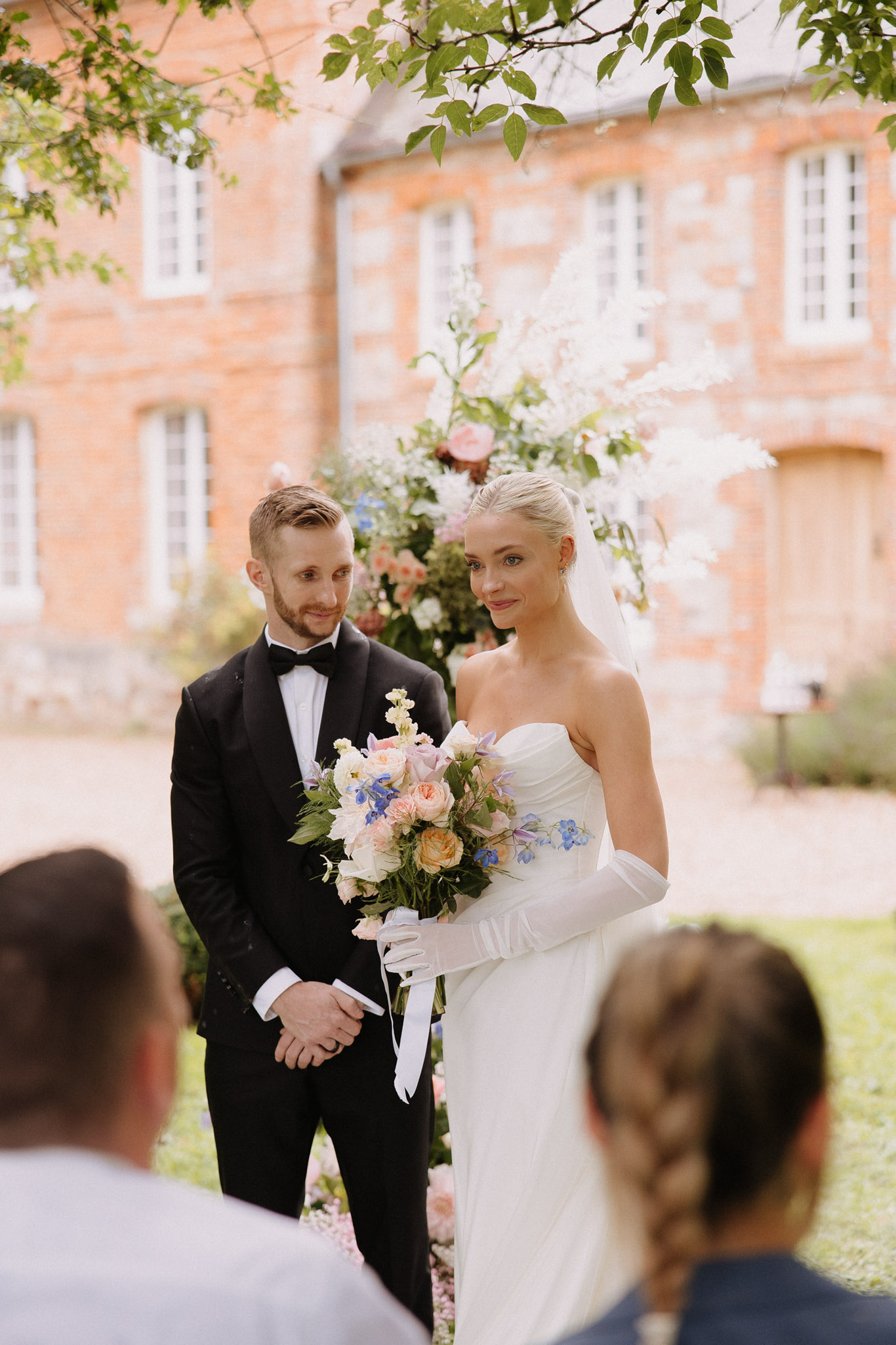 Couple at altar with garden-style peach rose and blue delphinium arch at red-brick chateau courtyard