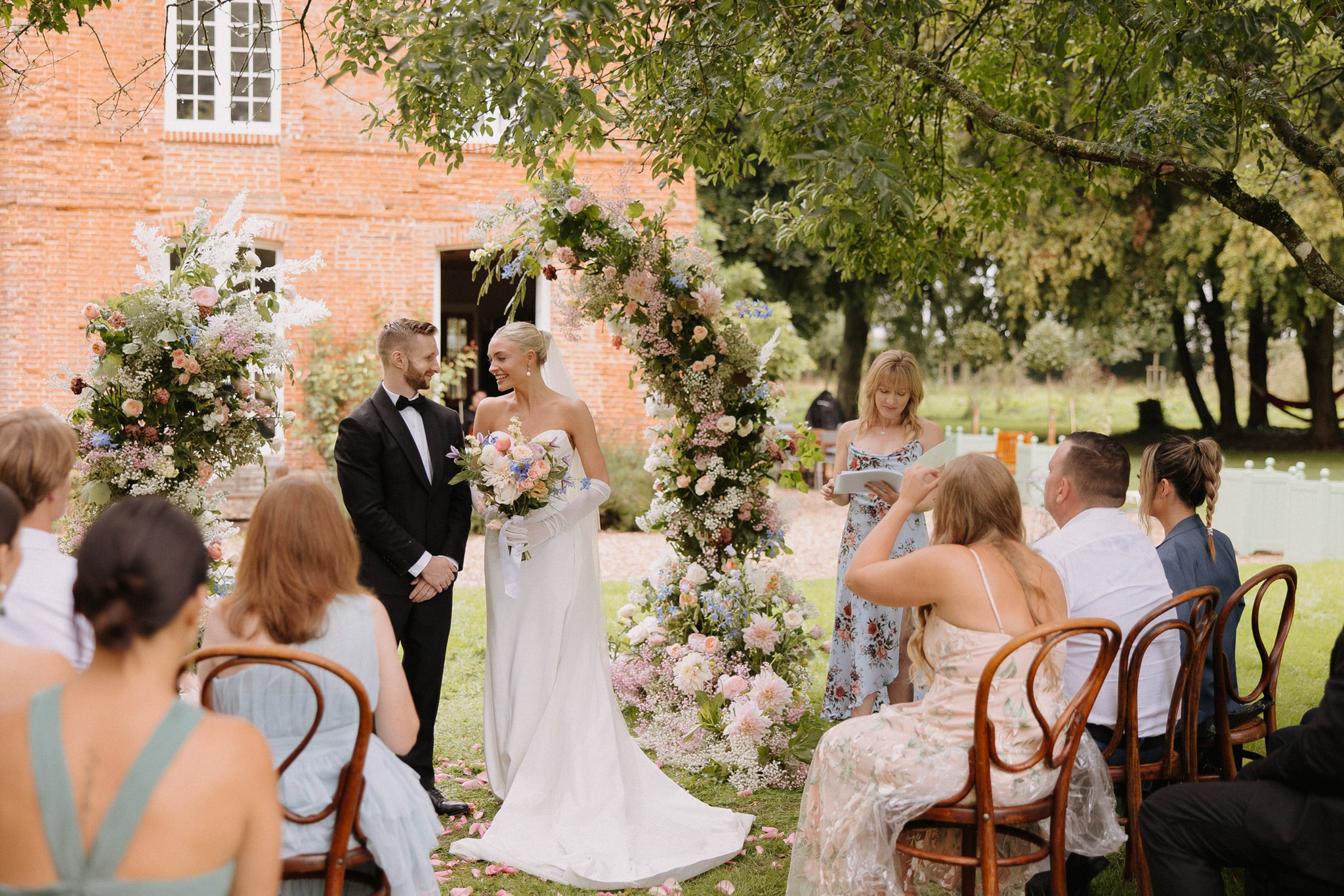 An outdoor wedding ceremony taking place in front of a red brick manor house, with the couple standing beneath a large circular floral arch densely covered in blush, peach, white, and periwinkle blue blooms including dahlias, peonies, astilbe, and gypsophila, with cascading greenery. The bride wears a white strapless fitted gown with a long train and elbow-length white gloves, holding a loose garden-style bouquet in peach, blush, and blue tones, while the groom wears a classic black tuxedo with bow tie; both are smiling and facing each other. A female officiant in a light blue floral dress reads from a tablet to their right, and approximately eight guests are seated on bentwood chairs arranged on the lawn, dressed in a mix of sage green, floral prints, and neutral tones. Loose flower petals are scattered along the aisle at the base of the arch, and the overall styling is romantic garden party with a soft pastel floral palette; wide shot taken from behind the seated guests.