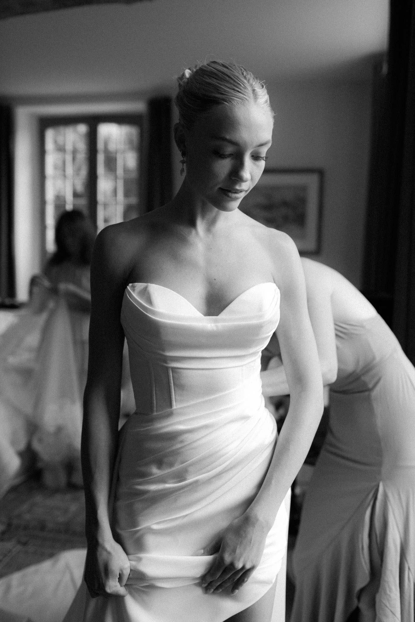 Black-and-white image of bride in strapless corset gown having dress adjusted during getting ready