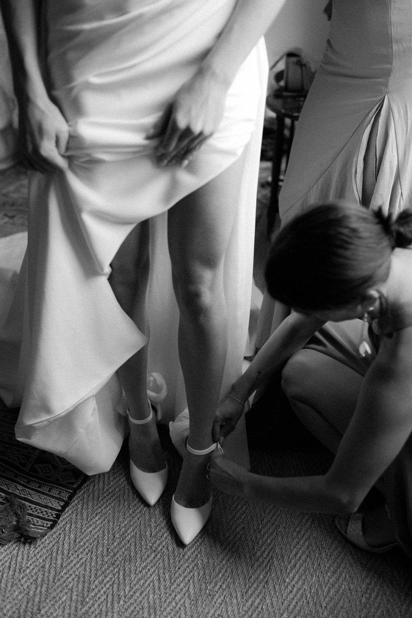 Bridesmaid kneeling to fasten ankle strap on bride's pointed-toe heels while bride lifts satin gown skirt