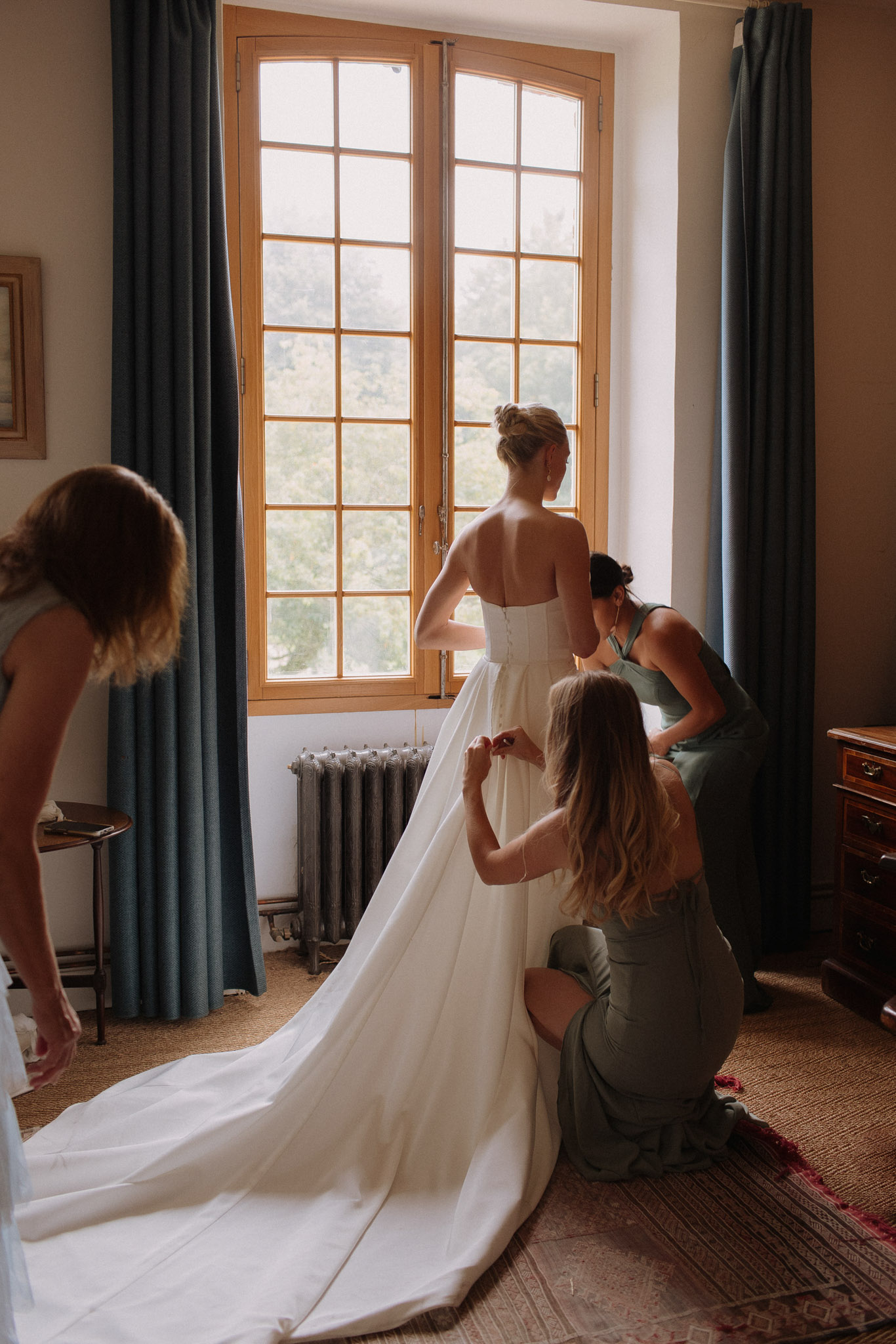 A getting-ready scene shot indoors, likely in a château bedroom or suite, with the bride standing at a large multi-pane wooden-framed window with slate blue curtains on either side. The bride is wearing a strapless white gown with a long cathedral-length train and has her hair up in a loose bun. Three bridesmaids in sage green satin dresses are assisting her: one kneels behind the bride fastening the back of the dress, another crouches to her right also helping with the buttons or lacing, and a third leans in from the left to arrange the train on the floor. The room features warm-toned walls, a cast-iron radiator, a patterned rug, and dark wood furniture. The shot is a medium-wide portrait taken from behind, with natural window light illuminating the scene.
