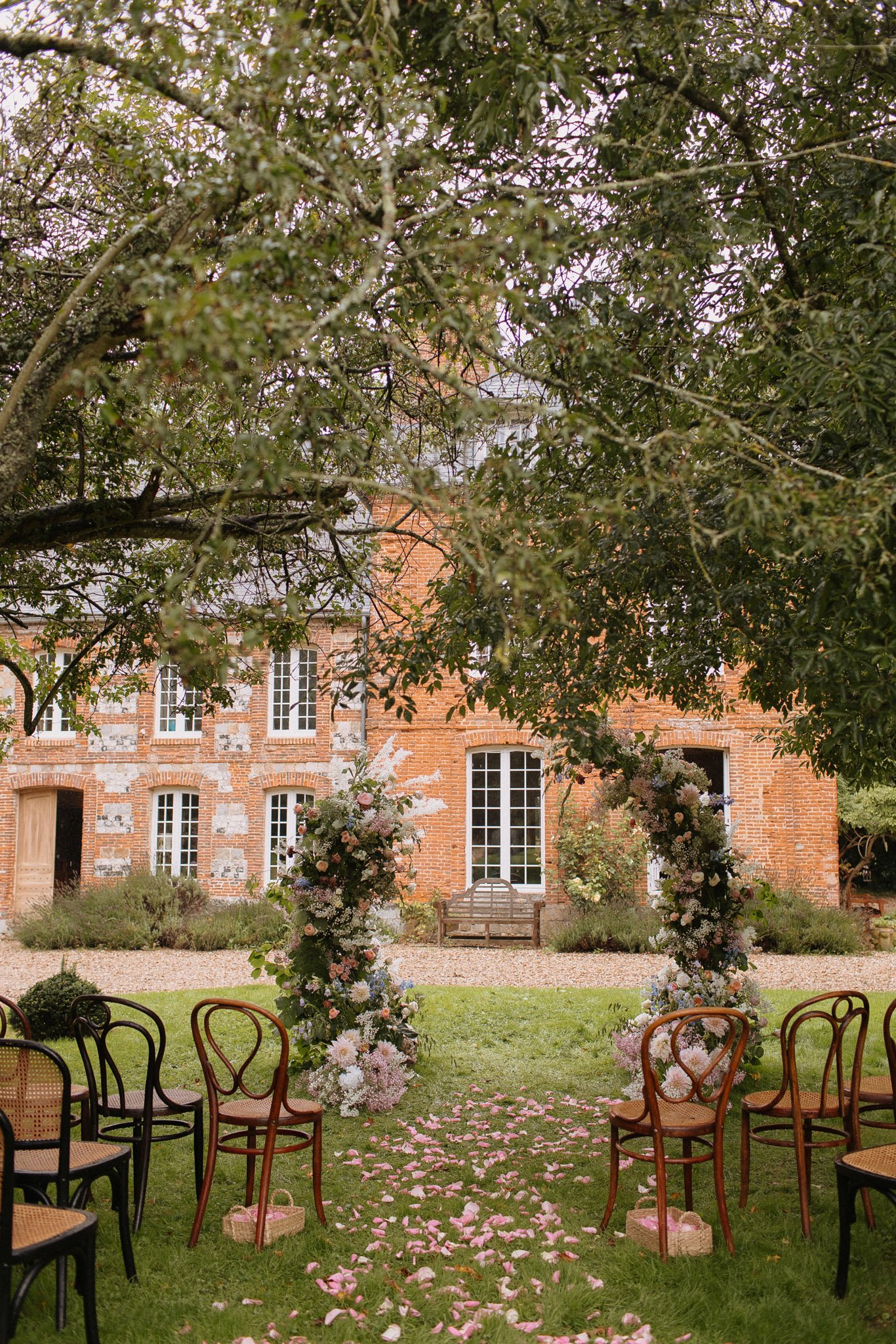 Garden ceremony setup with curved floral arch of peach, blush, and dusty blue blooms before red brick French manor