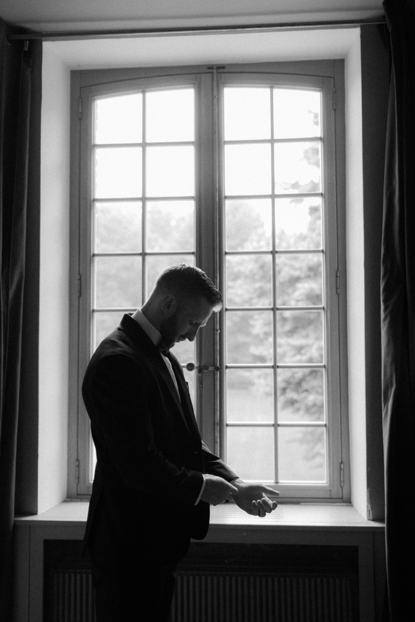 This black-and-white image shows a groom getting ready indoors, standing in front of a large arched multi-pane window that backlit him with bright natural light. He is adjusting his cufflinks or shirt sleeve, looking downward, and is dressed in a dark suit jacket with a dress shirt and tie. The strong contrast between the bright window and the darker interior creates a near-silhouette effect, with the groom's profile and hands remaining visible in the mid-tones. The window features classic French-style divided panes with arched tops, and a heavy curtain is partially visible on the right edge of the frame. The composition is a medium portrait shot taken from a slight distance, emphasizing the solitary, reflective mood of the pre-ceremony preparation moment.