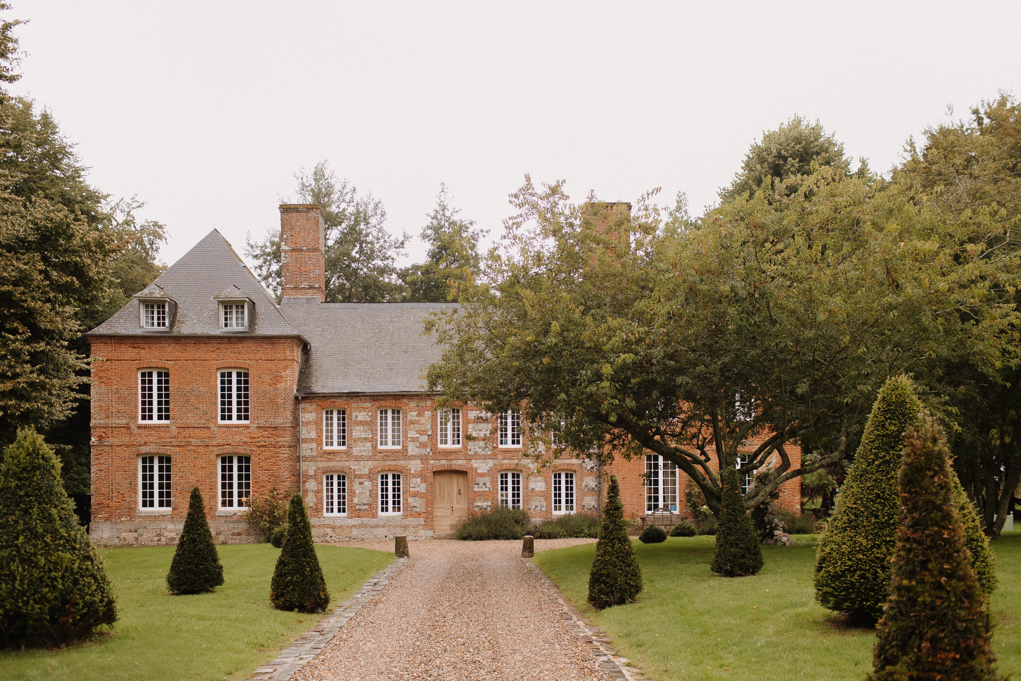 Exterior wide shot of a French manor house built in red brick and stone, featuring a grey slate roof with chimney stacks, white-framed multi-pane windows, and a central wooden door. A gravel driveway flanked by a symmetrical row of conical topiary shrubs leads directly to the entrance, creating a formal approach. The grounds include manicured lawn and mature trees framing the façade on both sides. No people are visible in the image. Potential venue feature image.