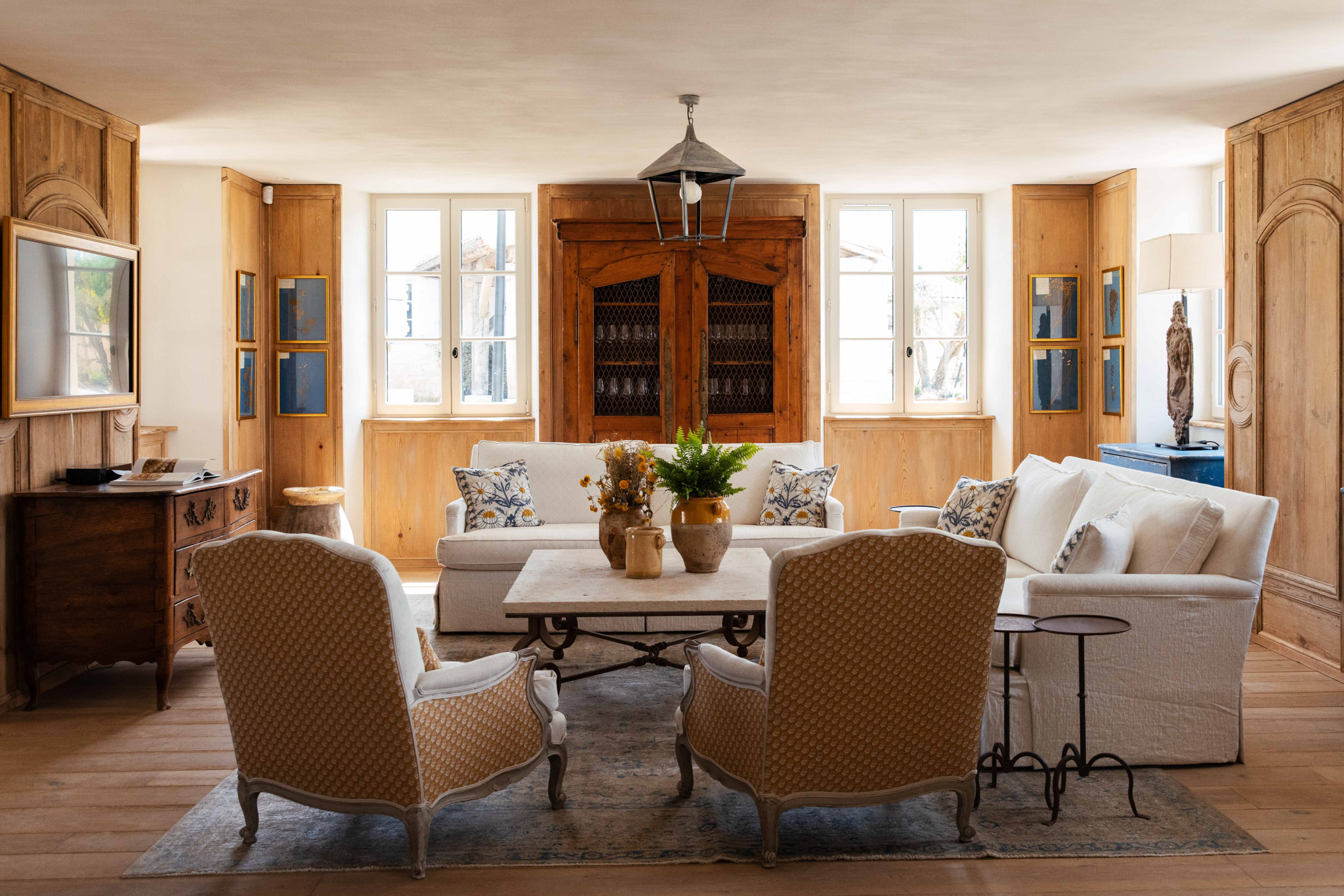 French country salon with honey wood paneling, linen sofas, Louis XV armchairs, and antique pine armoire