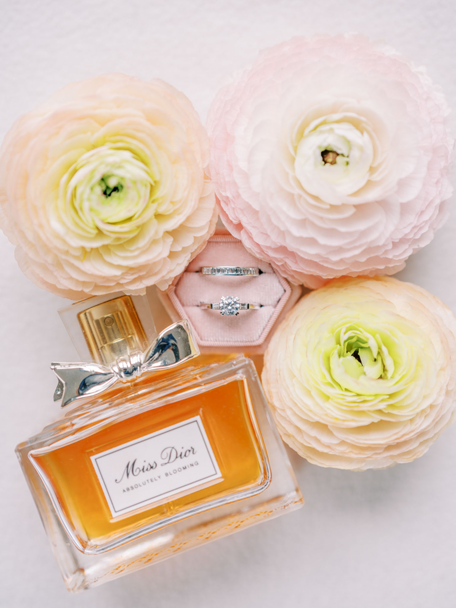 A flat lay detail shot of bridal accessories arranged on a white surface. A bottle of Miss Dior Absolutely Blooming perfume with a silver bow detail is positioned in the lower left, alongside a blush pink hexagonal velvet ring box holding two rings — a round solitaire diamond engagement ring and a diamond eternity band. Three large ranunculus blooms in blush pink and cream-green surround the items, framing the composition. The overall palette is soft blush, cream, and gold, consistent with a classic, feminine styling approach.
