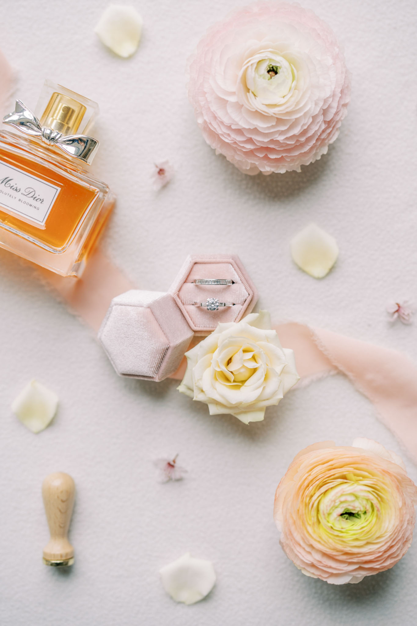 A flat lay detail shot arranged on a white surface featuring two wedding bands — a diamond solitaire engagement ring and a plain band — displayed in an open hexagonal blush velvet ring box. Surrounding the ring box are a bottle of Miss Dior Absolutely Blooming perfume, a cream garden rose, two blush and peach ranunculus blooms, scattered cream and blush petals, a small wooden wax seal stamp, and a length of sheer blush ribbon. The overall styling palette is soft blush, cream, and peach, consistent with a romantic, classic French wedding aesthetic.
