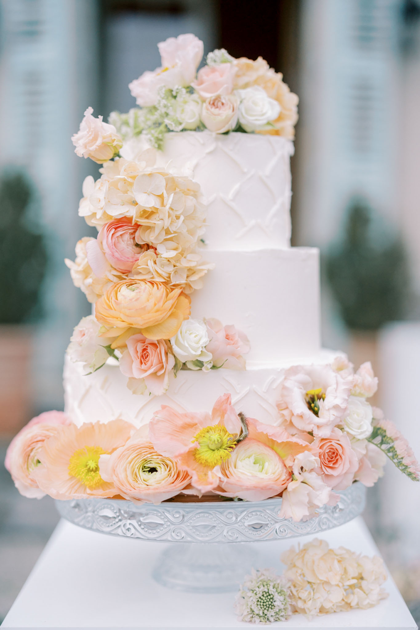 Four-tier white quilted wedding cake with cascading peach ranunculus blush roses cream hydrangeas on silver stand