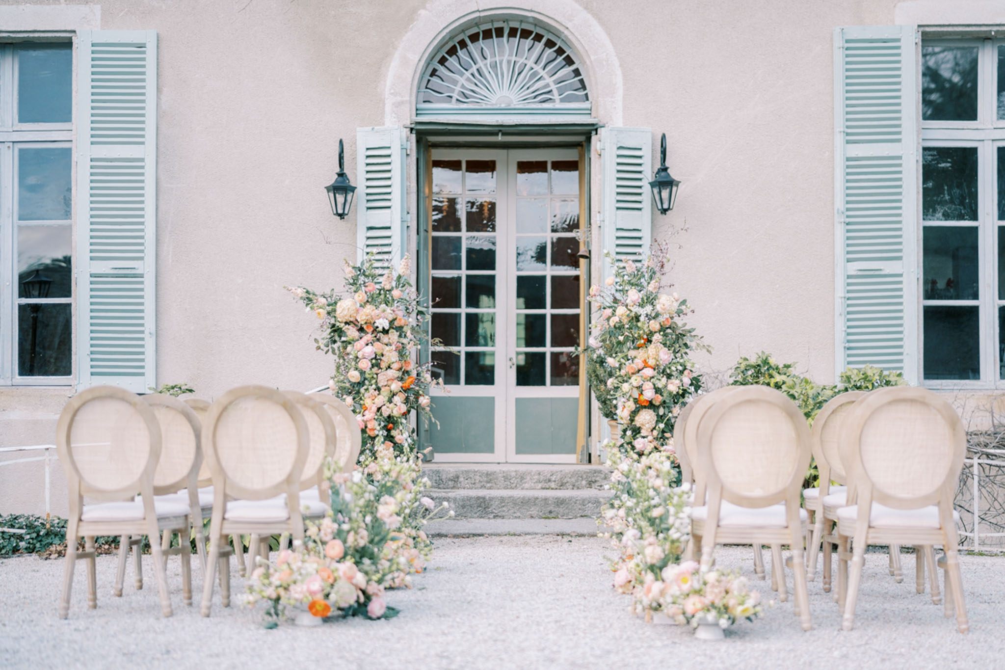 An outdoor wedding ceremony setup photographed in a wide shot, positioned in front of a French château facade with sage green shutters, arched fanlight door, and pale stone rendering. Two rows of natural linen-upholstered Louis XVI-style oval-back chairs are arranged on a gravel courtyard, divided by a central aisle. Tall floral columns flank the château entrance door, composed of blush, peach, coral, and ivory garden roses, ranunculus, and trailing greenery. Matching low floral clusters are placed at the base of the aisle entrance, repeating the same peach, blush, and orange tonal palette with abundant green foliage. The overall styling is classic French with a soft warm floral palette against the cool sage architectural details. No guests or wedding party are present in the image. Potential venue feature image.