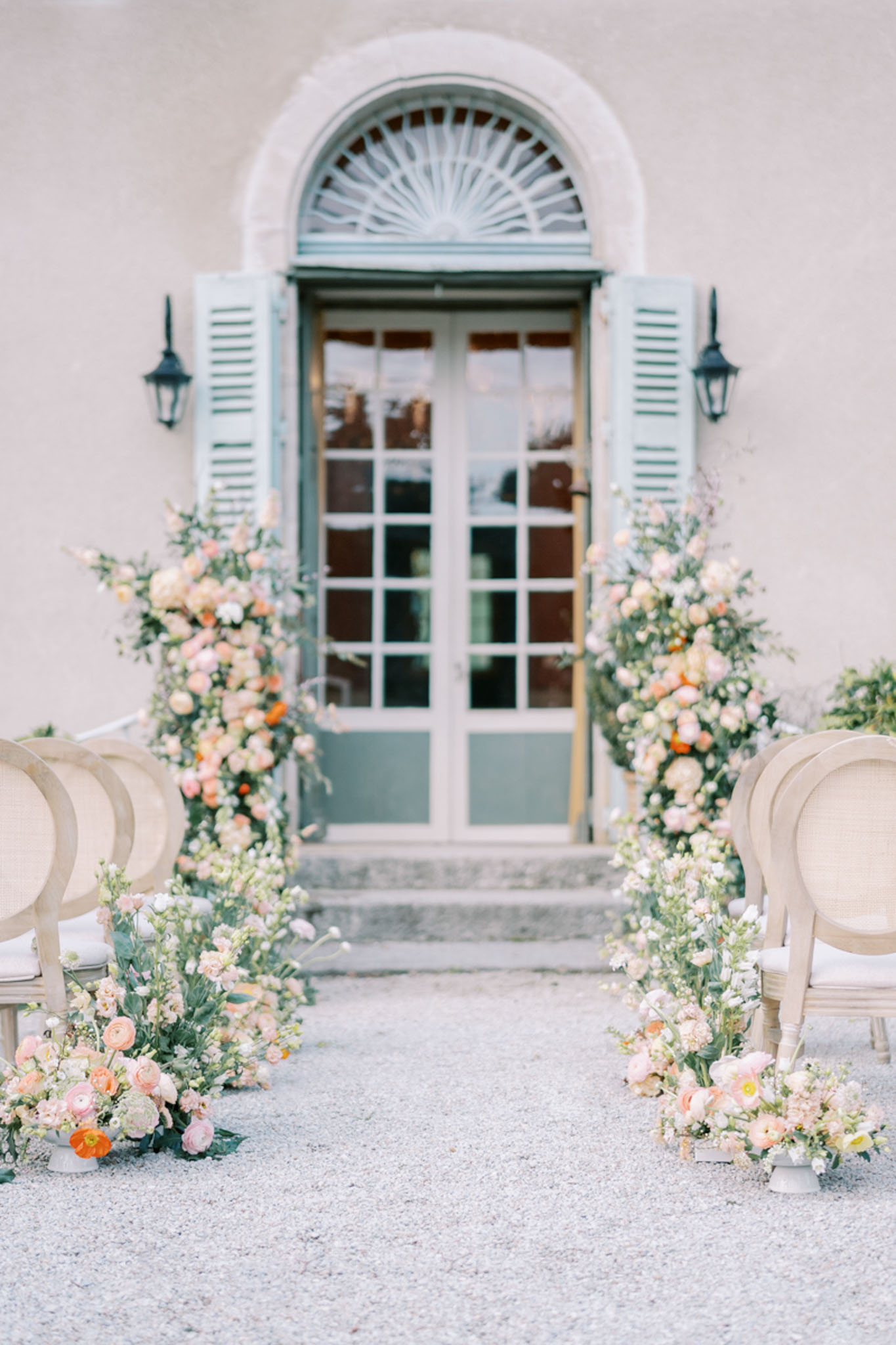 An outdoor wedding ceremony setup photographed from the aisle looking toward a French château entrance, with no people present. The ceremony aisle is flanked by rows of Louis XVI-style chairs in natural linen upholstery on a gravel surface. Two tall floral column installations frame the arched doorway, composed of blush pink ranunculus, peach garden roses, white stock, orange poppy accents, and trailing greenery. Smaller low floral arrangements in white ceramic vessels are placed at the foot of each column and along the front chair rows, using the same blush, peach, and cream palette. The backdrop is a pale stone façade with a set of sage green shuttered French doors topped by a fanlight arch and flanked by black iron lanterns. The overall styling is classic French with a soft peach and blush floral palette. Wide shot, centered composition. Potential venue feature image.