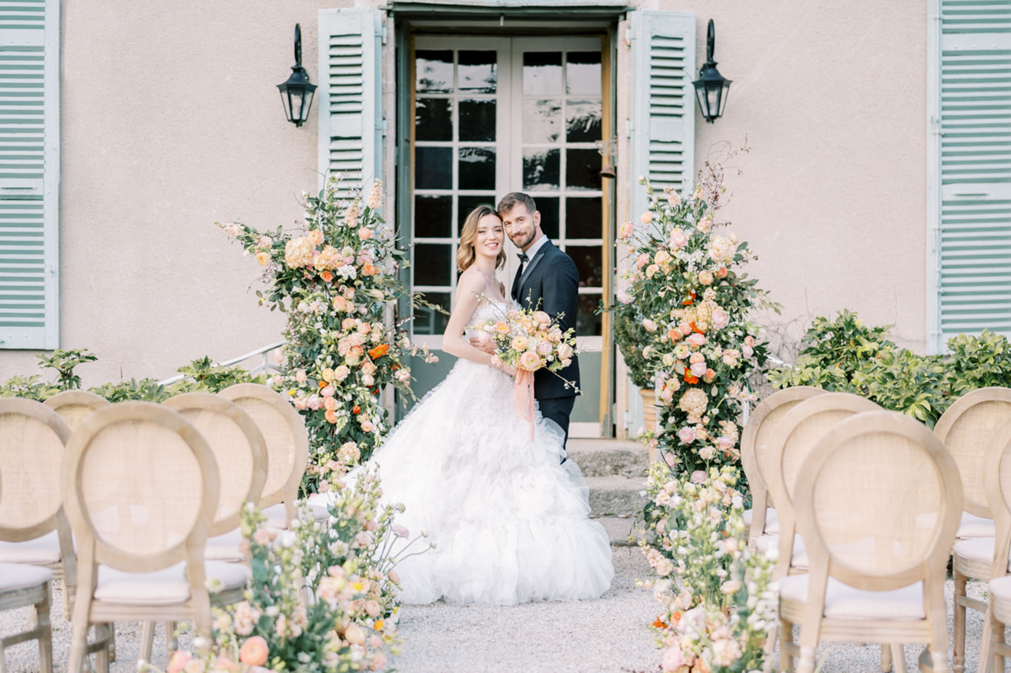 Bride and groom at outdoor altar flanked by tall peach and blush floral columns before French manor with green shutters
