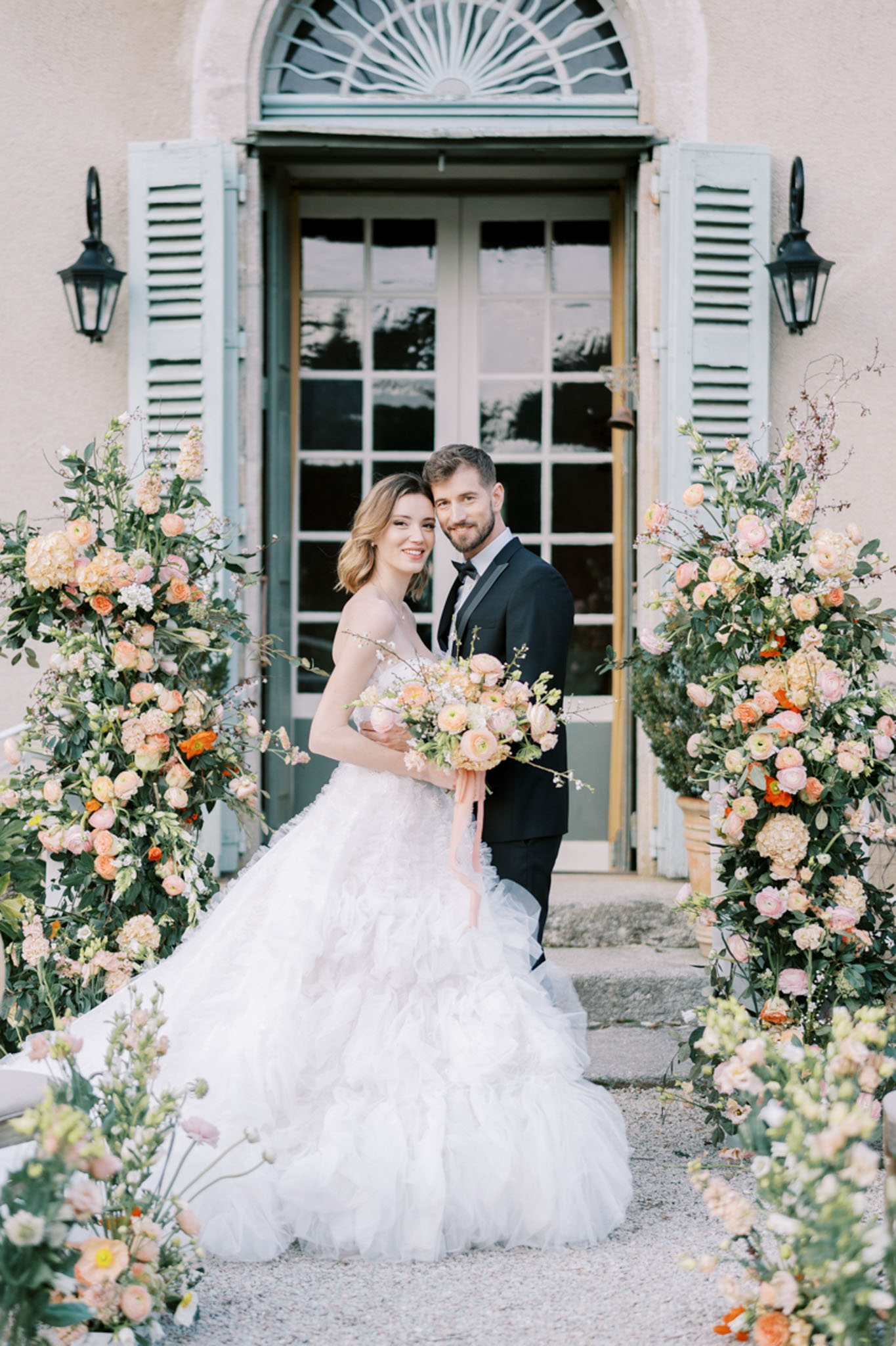 A couple poses for a portrait outdoors in front of a French château entrance featuring sage green shutters, a fanlight transom window, and black iron wall lanterns. The bride wears a strapless white ruffled tulle ball gown and holds a bouquet of peach ranunculus, blush garden roses, and cream blooms tied with a trailing blush silk ribbon. The groom is dressed in a black tuxedo with a black bow tie. They are framed on both sides by tall, lush floral column arrangements in a peach, blush, coral, and orange palette featuring ranunculus, garden roses, hydrangea, and mixed greenery, with additional low floral clusters at ground level. The overall styling is classic French with a warm, garden-inspired floral palette. Medium portrait shot from approximately knee height.
