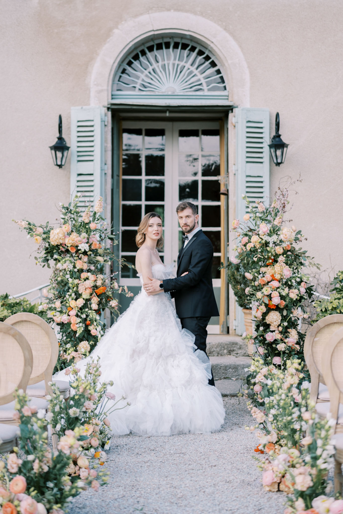 A couple portrait taken outdoors in front of a French manor or château facade featuring a pale blush-rendered wall, a stone-framed arched doorway with blue-grey shutters, and wrought-iron wall lanterns. The bride wears a strapless, tiered ruffled ball gown in white, and the groom wears a black tuxedo with a bow tie; both face the camera while she rests her hand on his arm. They are framed by two large freestanding floral column arrangements in peach, coral, orange, blush pink, and cream tones featuring ranunculus, hydrangeas, snapdragons, and lush greenery. The ceremony aisle is lined with additional low floral arrangements in the same palette, and natural cane-back chairs are partially visible on either side. The overall styling is classic French with a warm, garden-party floral palette. Medium portrait shot. Potential venue feature image.