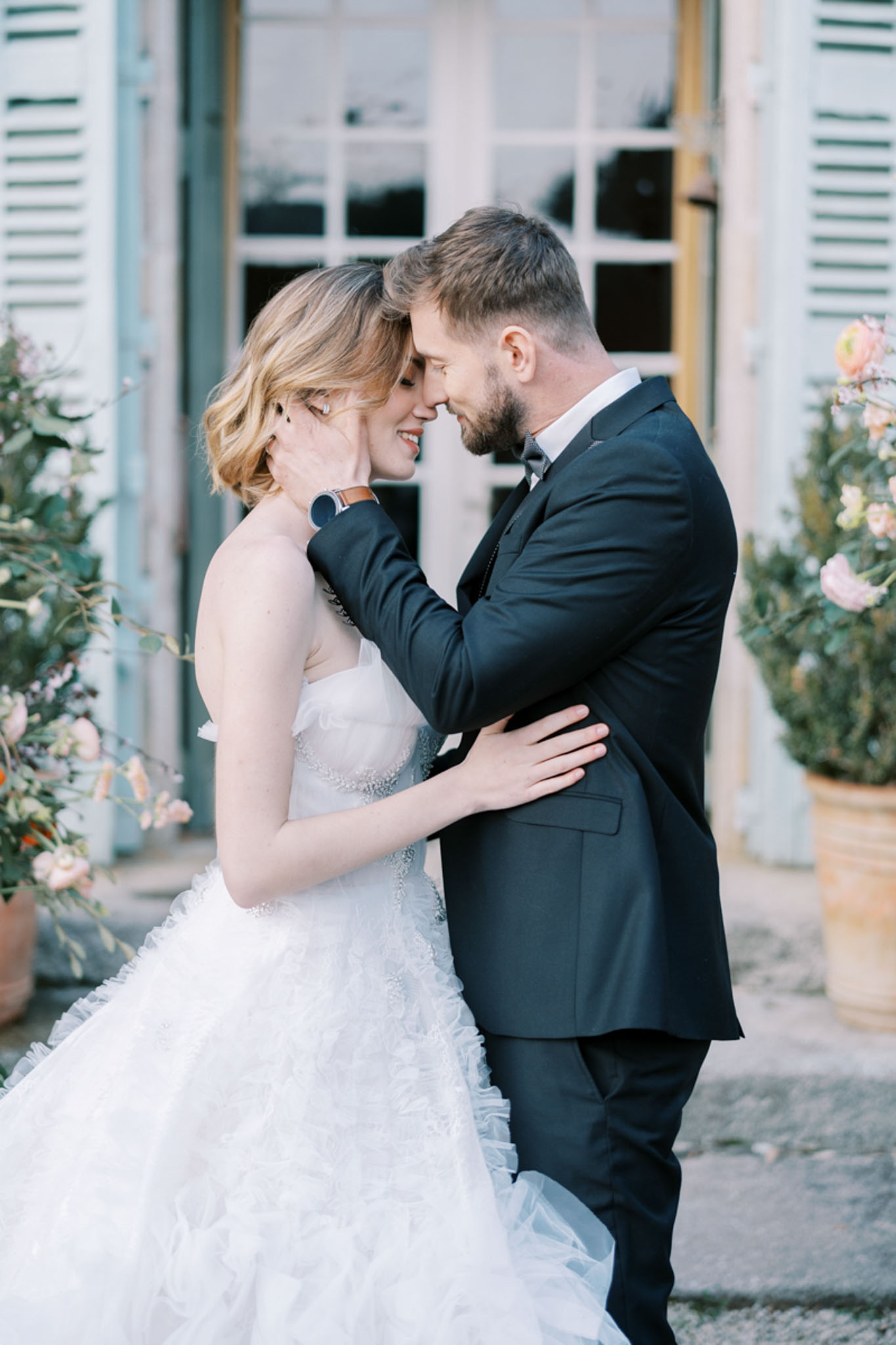 A couples portrait showing the bride and groom standing close together with foreheads nearly touching, photographed from a medium distance outdoors in front of a French-style building with pale blue-grey shutters and tall glass doors. The bride wears a strapless white ballgown with a ruffled layered tulle skirt and a beaded waistband, with short wavy blonde hair; the groom wears a dark navy suit with a grey bow tie and a leather-strap watch. Large terracotta pots filled with blush pink roses and trailing greenery flank the doorway on both sides, adding a soft romantic palette to the background. The overall styling aesthetic is classic French château with a soft, airy feel.