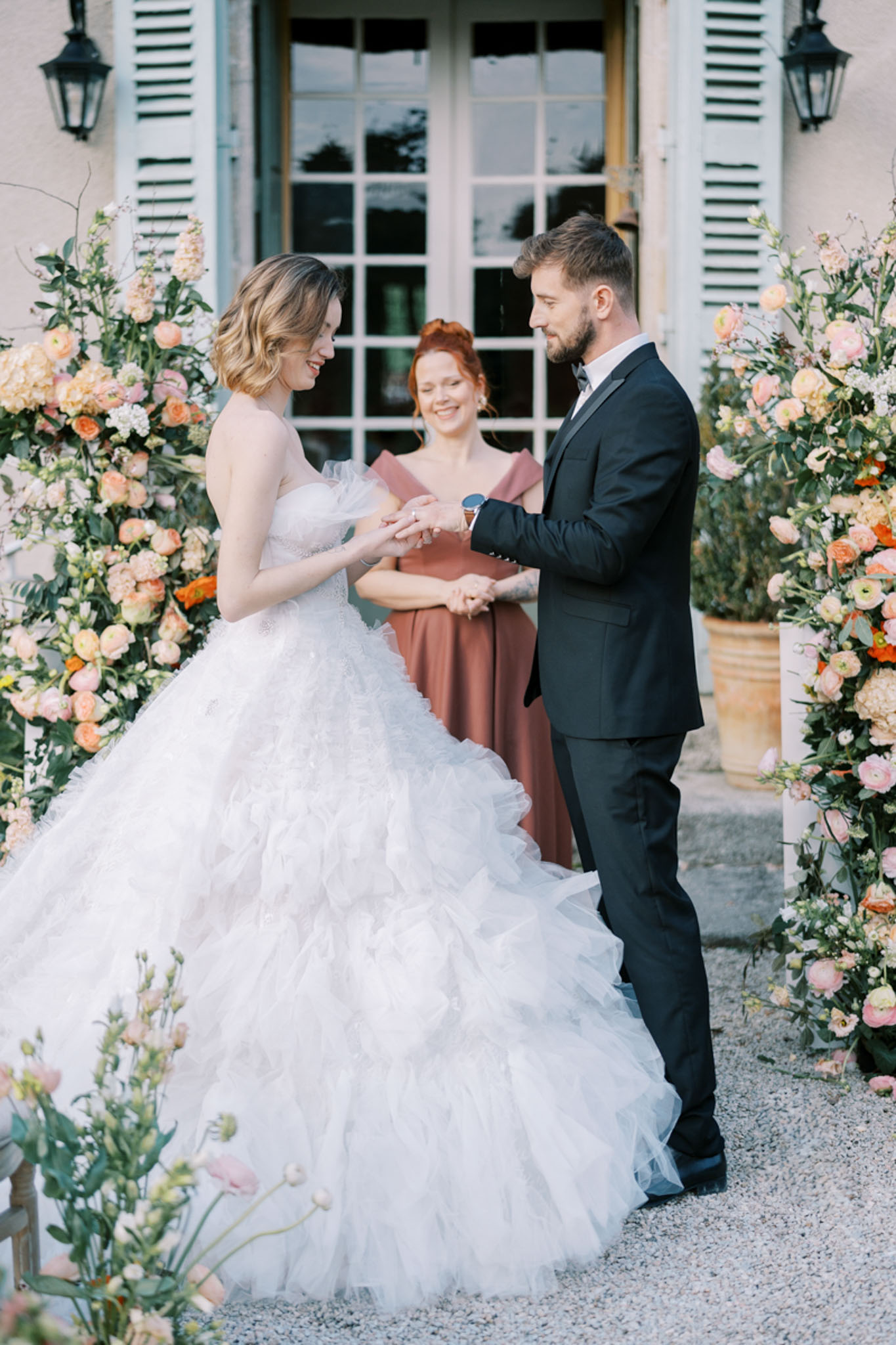The ring exchange moment of an outdoor wedding ceremony taking place in front of a French château facade with pale blue shutters and tall French windows. The bride wears a strapless ivory ballgown with a heavily layered, ruffled tulle skirt and a fitted lace bodice, while the groom wears a dark navy slim-fit suit with a black bow tie. A female officiant stands between them wearing a dusty rose off-the-shoulder gown and holds a small book. Two large floral arrangements frame the couple on either side, featuring peach ranunculus, blush garden roses, orange blooms, and lush greenery in terracotta urns. The ceremony takes place on a gravel surface, and the overall decor palette is peach, blush, and coral with a classic French-country aesthetic. The shot is a medium full-length portrait capturing all three figures.