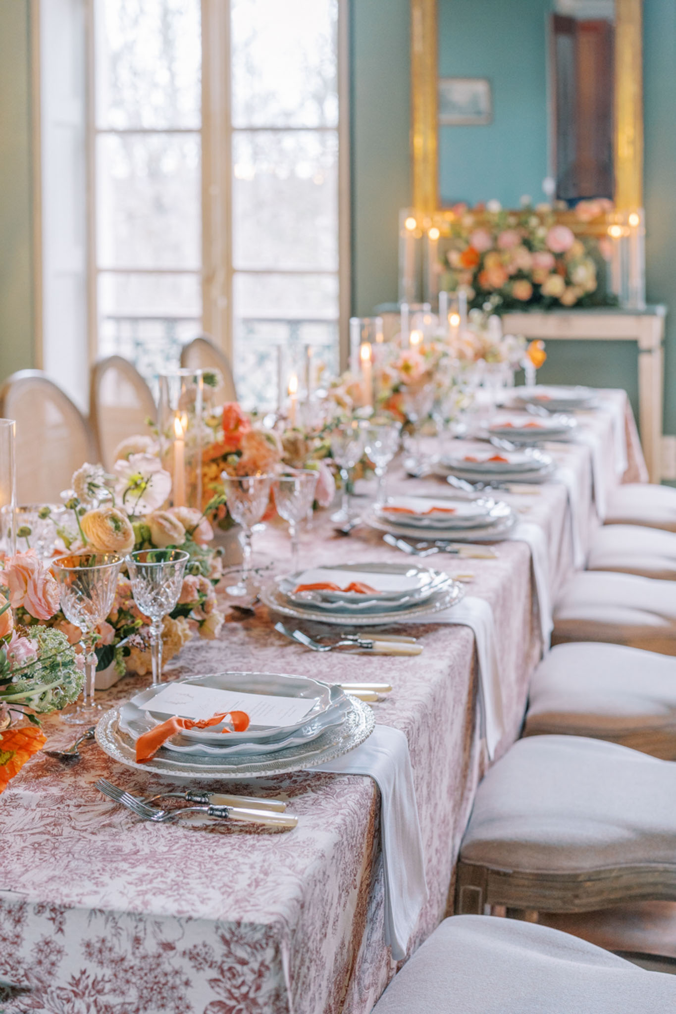 A reception tablescape detail shot taken indoors in a room with sage green walls, tall French windows, and a gold-framed mirror above a fireplace mantel. The long dining table is dressed in a blush pink toile de Jouy patterned tablecloth and set for approximately ten guests, with upholstered Louis XVI-style chairs in cream and natural linen. Each place setting features a silver charger plate layered with a scallop-edged grey dinner plate, a white menu card, and a folded burnt orange ribbon accent. Crystal cut-glass wine and water goblets, bone-handled flatware, and silver cutlery complete each setting. A lush floral runner runs the length of the table featuring peach garden roses, coral ranunculus, blush lisianthus, orange blooms, and green foliage, interspersed with tall clear glass taper candle holders with lit candles. A matching floral arrangement sits on the fireplace mantel in the background, flanked by additional candlelight. The overall palette combines blush, coral, burnt orange, and sage green in a classic French interior style.