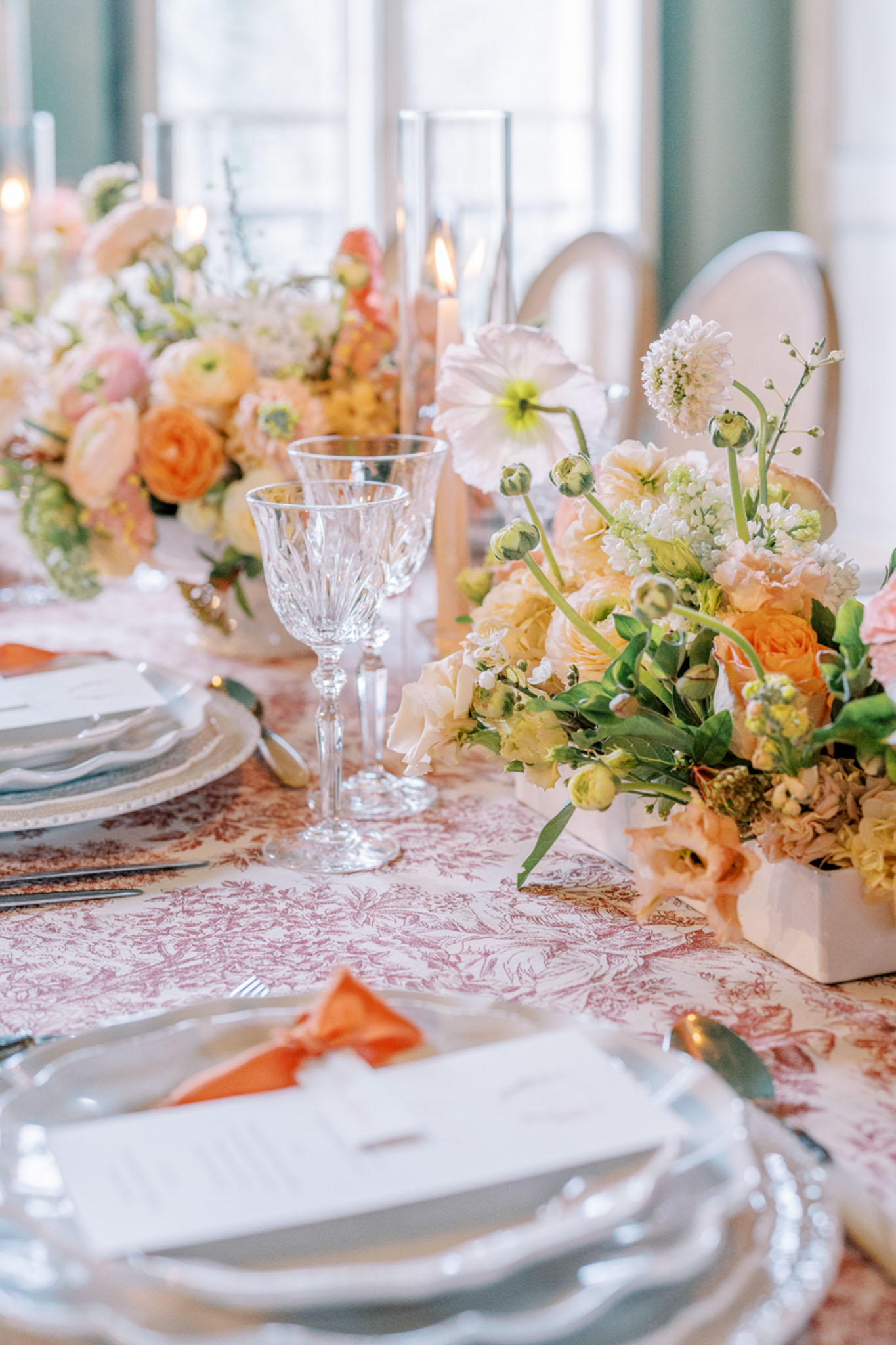 Toile tablecloth with peach roses, orange ranunculus, coral napkins, and scallop-edged charger plates