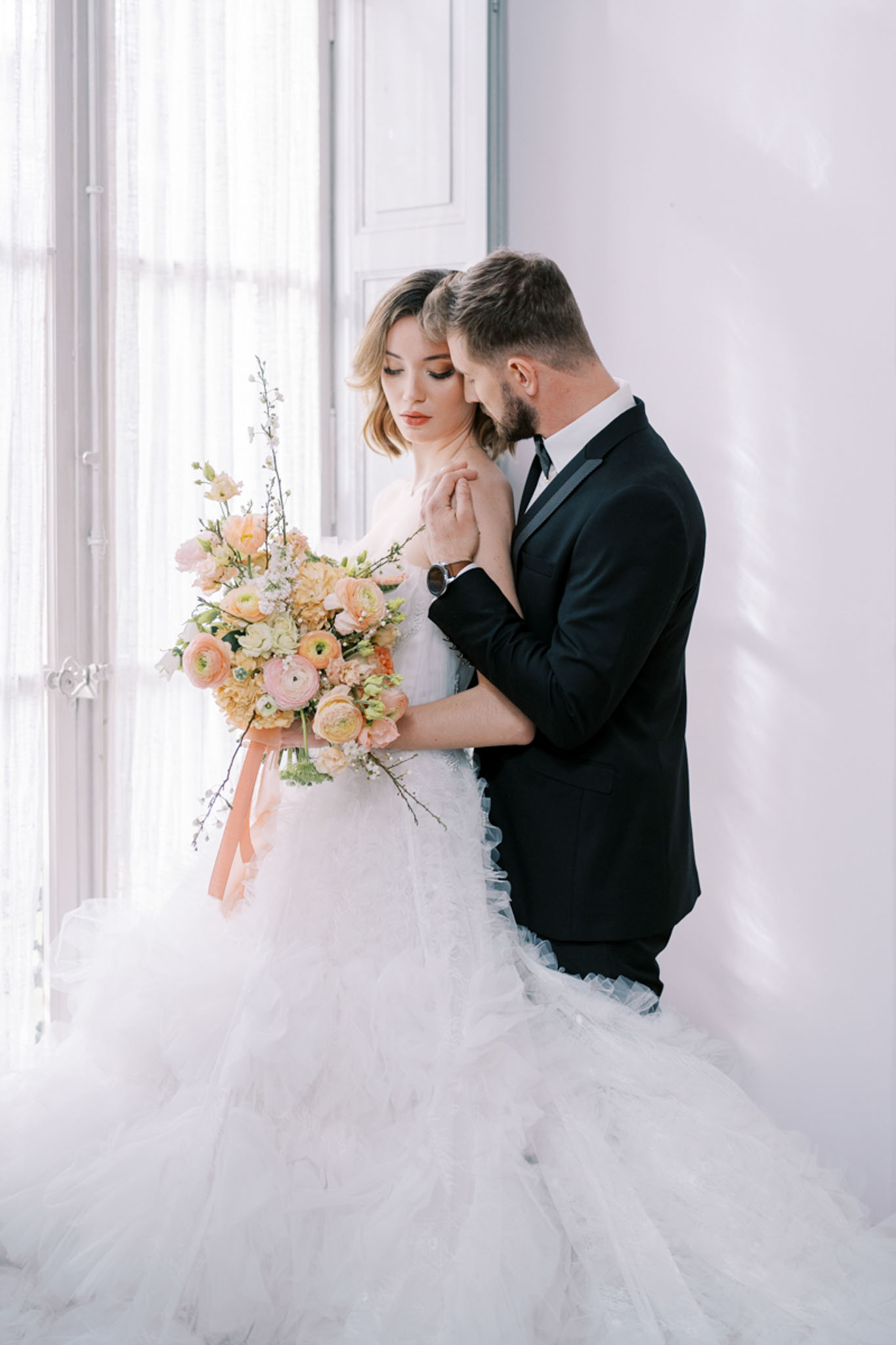 A couple portrait shot indoors against a white wall beside a large window dressed with sheer white curtains. The bride wears a white ballgown with a voluminous layered tulle skirt and holds a loose, garden-style bouquet composed of peach and blush ranunculus, cream lisianthus, and delicate white branching blooms, tied with a peach satin ribbon. The groom stands behind her wearing a black tuxedo jacket with a black tie and white dress shirt, his arms wrapped around her as he leans his head toward hers. The overall styling is modern classic with a soft peach and white color palette. The image is a medium portrait shot with soft natural backlight from the window.