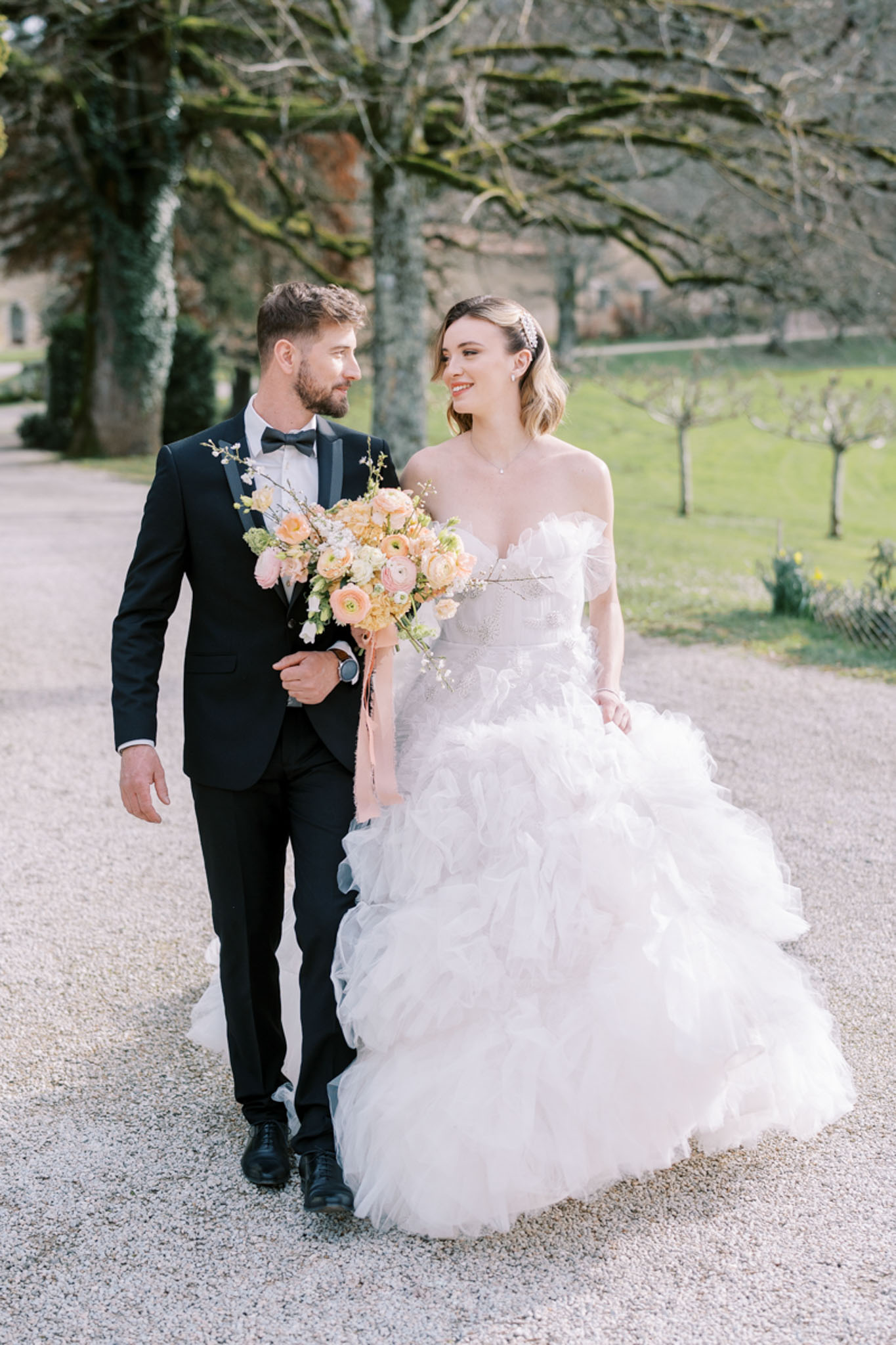 Bride in off-shoulder ruffle tulle ballgown and groom in black tuxedo walking on gravel path with peach bouquet