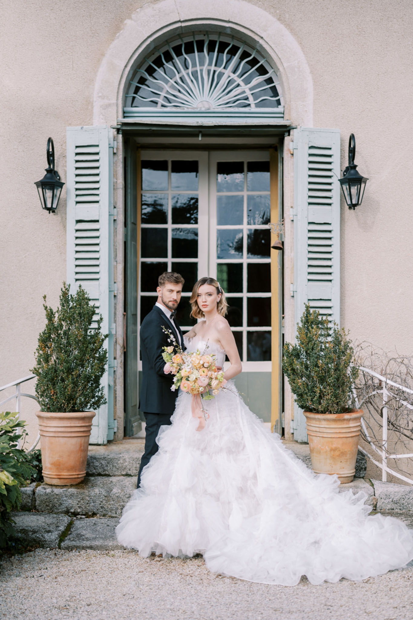 Bride in tiered tulle ballgown with peach bouquet and groom in navy suit before chateau entrance with topiary