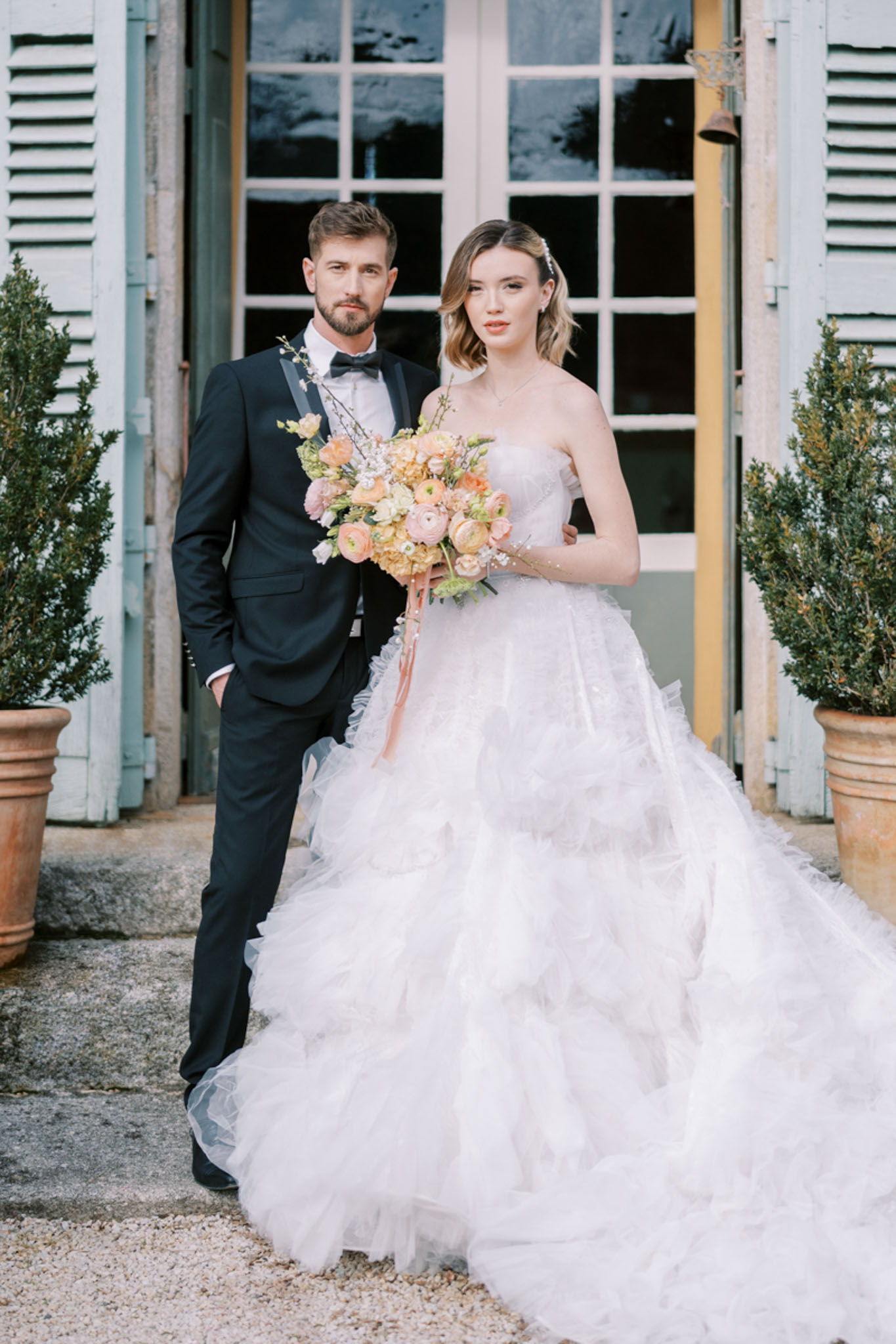 A couple portrait taken outdoors in front of a French château entrance featuring pale blue-grey shutters, large French windows, and terracotta planters with topiary shrubs on either side. The groom wears a navy tuxedo with black satin lapels, a white dress shirt, and a black bow tie, while the bride wears a strapless ivory ball gown with a heavily layered and ruffled tulle skirt. She holds a rounded bouquet composed of peach and coral ranunculus, blush peonies, soft yellow hydrangea, white gypsophila, and trailing peach ribbon. The styling is classic French château with a formal, polished aesthetic. The shot is a mid-length portrait with both subjects facing the camera directly.