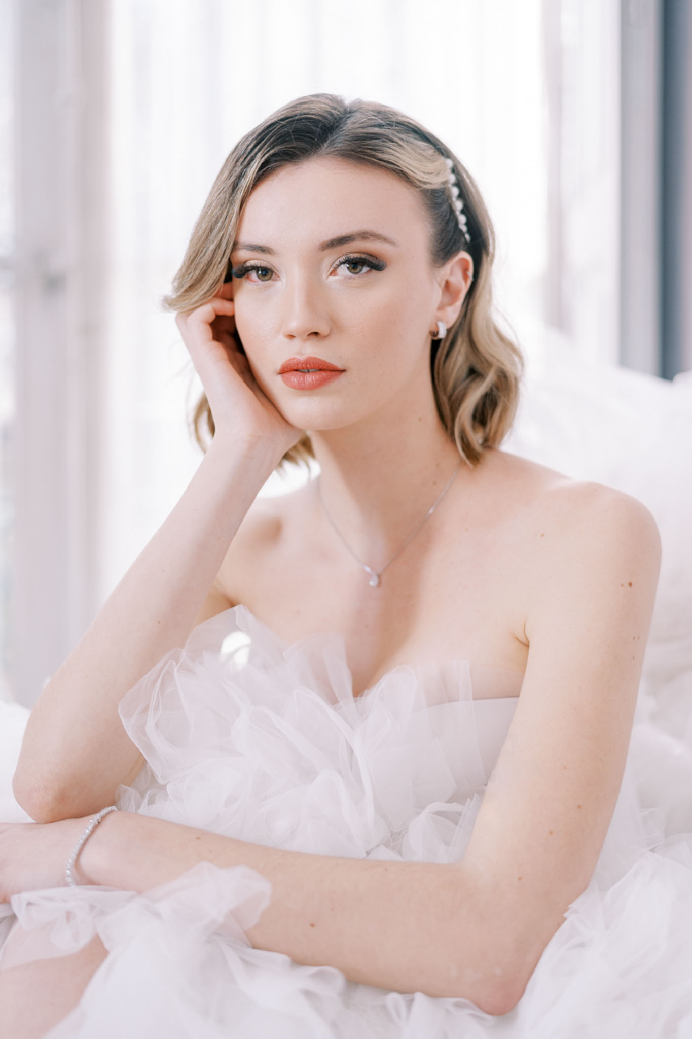 Bride in a strapless white tulle ballgown with pearl headband posing by a bright window during getting ready
