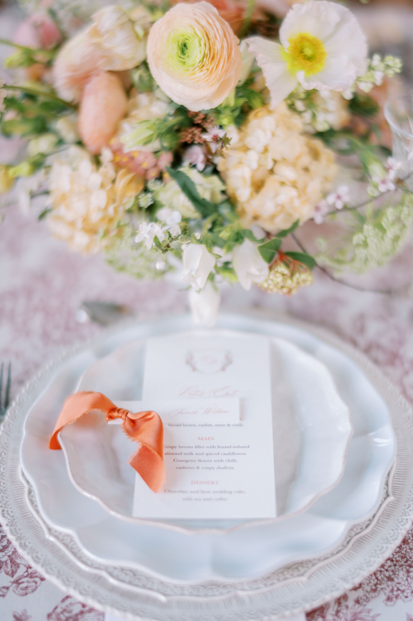 A close-up detail shot of a wedding reception place setting featuring a white scallop-edged charger plate layered with a pale blue ceramic dinner plate, on which rests a printed menu card tied with a burnt orange satin ribbon bow. The menu card has coral-colored text and is printed with starter, main, and dessert courses. The table linen beneath is a dusty rose toile fabric. In the soft-focus background, a floral centerpiece is visible, composed of blush and cream ranunculus, ivory hydrangea, white poppies, peach tulips, and delicate small white blooms with green foliage, arranged in a loose, garden-style design. The overall decor palette combines cream, peach, blush, and burnt orange accents in a romantic, classic style.