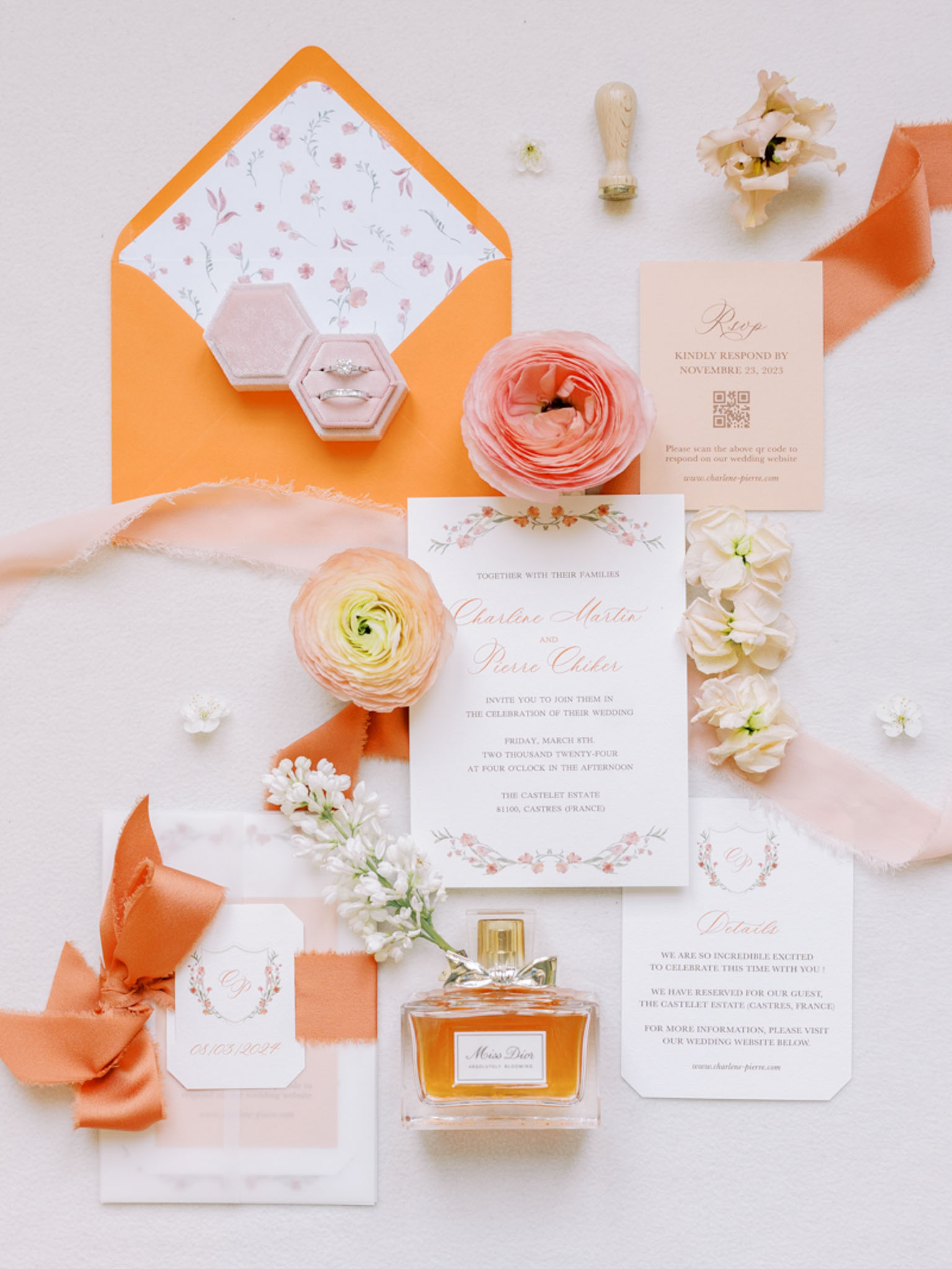 Wedding stationery flat lay with coral floral invitations, velvet ring box, Miss Dior perfume, and ranunculus