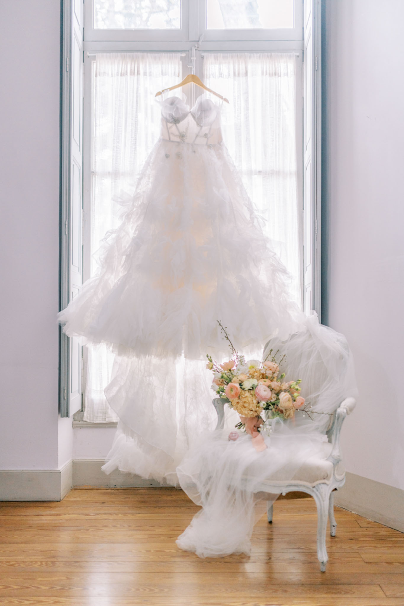 Ivory ruffled ballgown on hanger at French window with blush ranunculus bouquet on Louis XV chair