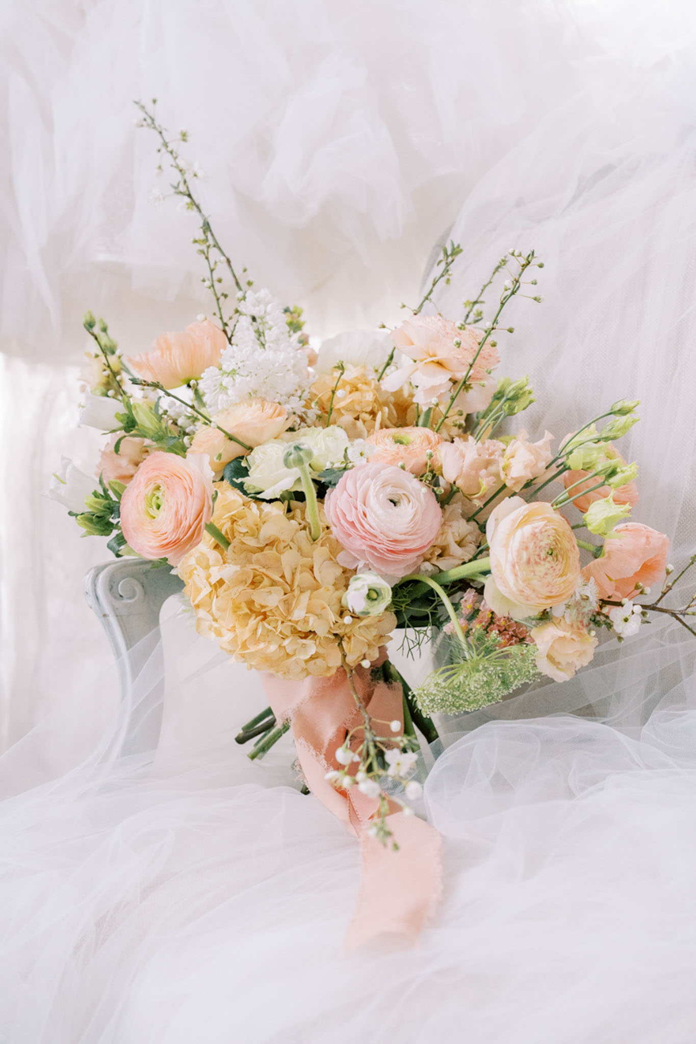 Bridal bouquet of peach ranunculus and blush flowers with pink ribbon resting on white tulle