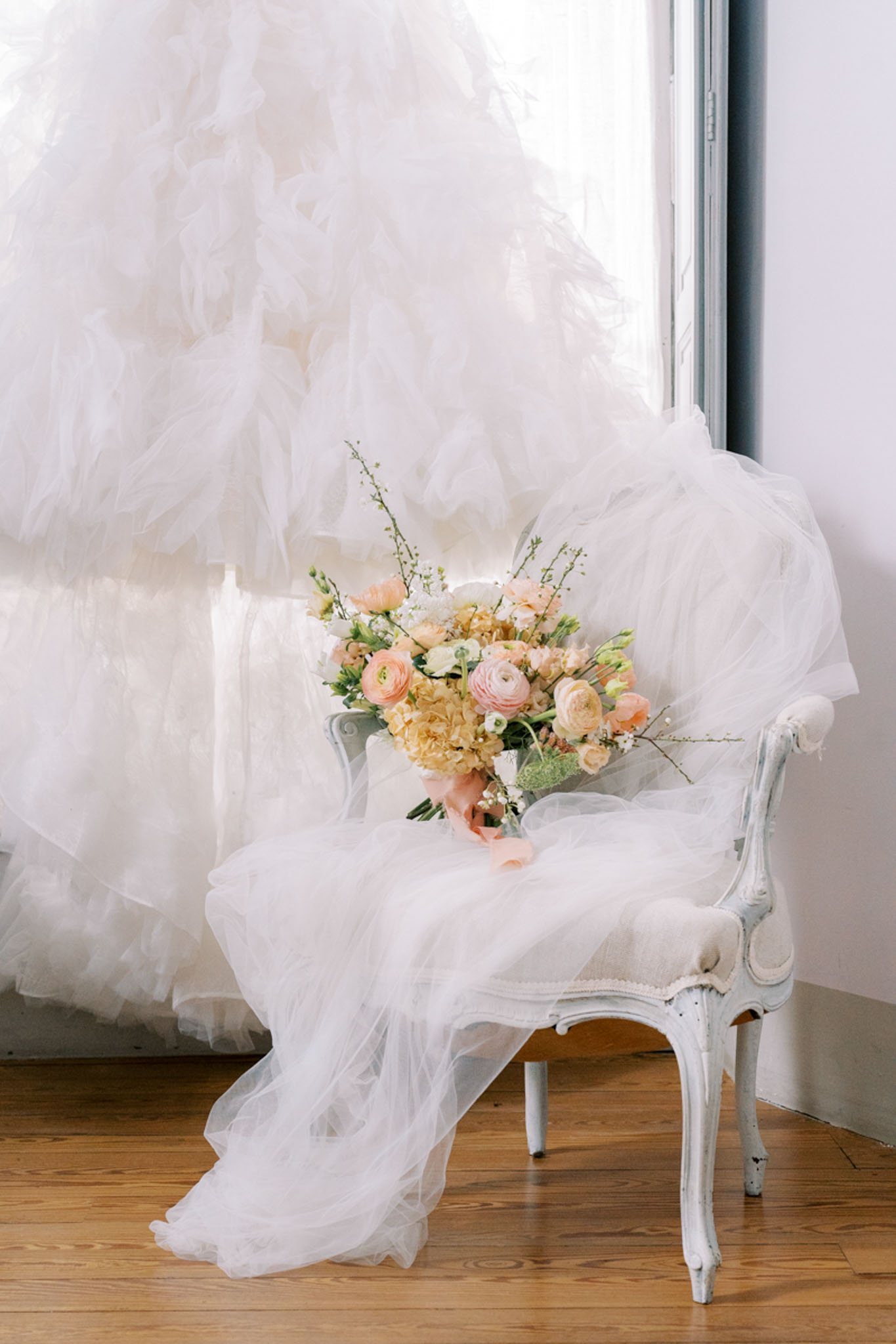 White ruffled tulle ballgown draped on painted armchair with blush ranunculus and peach rose bouquet