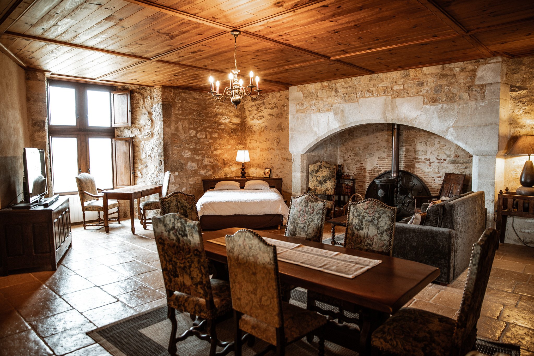 Interior wide shot of a large room within what appears to be a French château or historic manor, showing accommodation available at the venue. The room features exposed limestone walls, a wood-panelled coffered ceiling, and terracotta stone tile flooring. A substantial arched stone fireplace with a black wood-burning stove dominates the right side of the room. Furnishings include a dark wood dining table surrounded by six tapestry-upholstered chairs with brass nail-head trim, a double bed with white linens, a dark grey velvet sofa, and several antique-style side chairs. Lighting comes from a brass candle-style chandelier, a traditional table lamp, and natural light through a wooden-shuttered window. The overall decor style is classic French rustic with period-appropriate furniture throughout. Potential venue feature image.